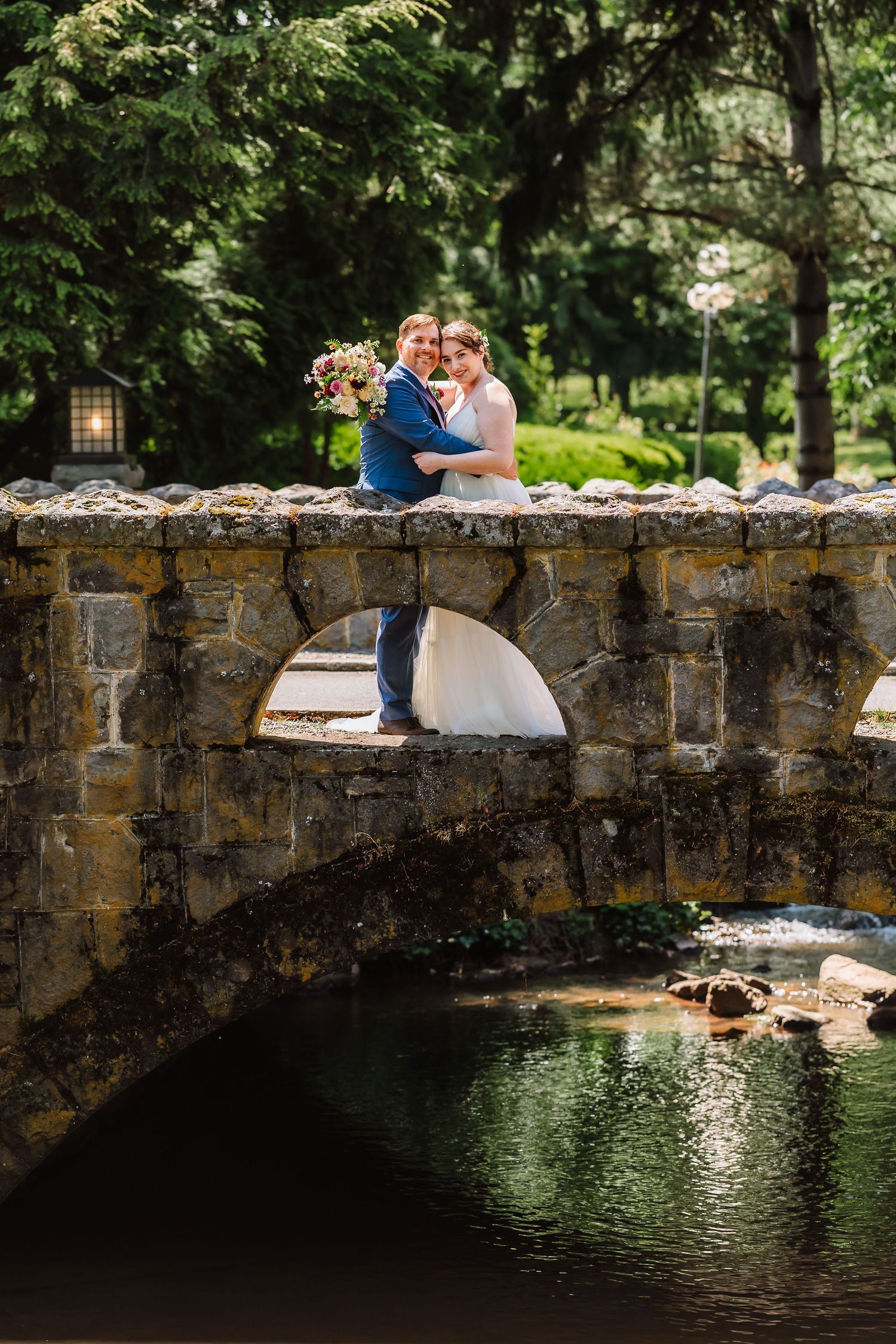 Newlyweds embracing on a rustic stone bridge over a stream in a lush garden setting.