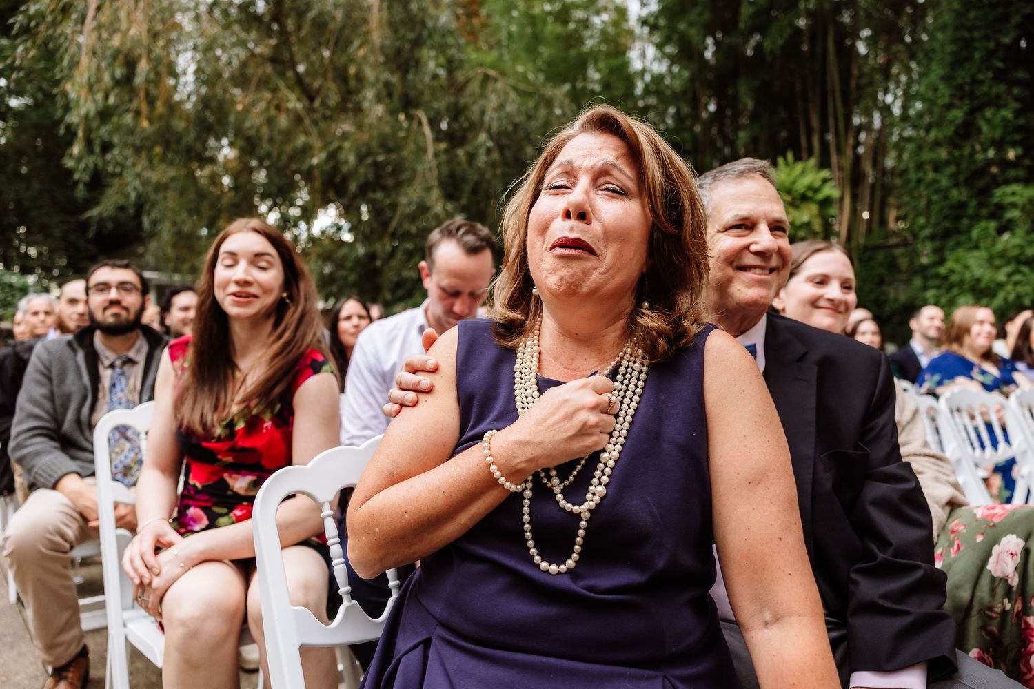 Emotional woman in purple dress touches her heart during outdoor wedding ceremony.