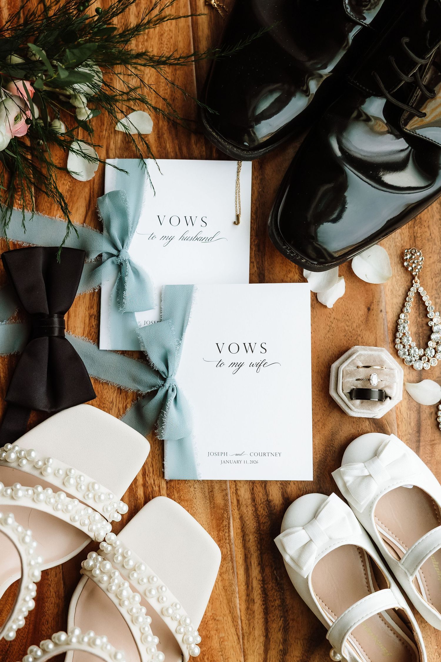 Wedding vow booklets with blue ribbon bows surrounded by bridal shoes, groom dress shoes, jewelry, and floral accents on wood.
