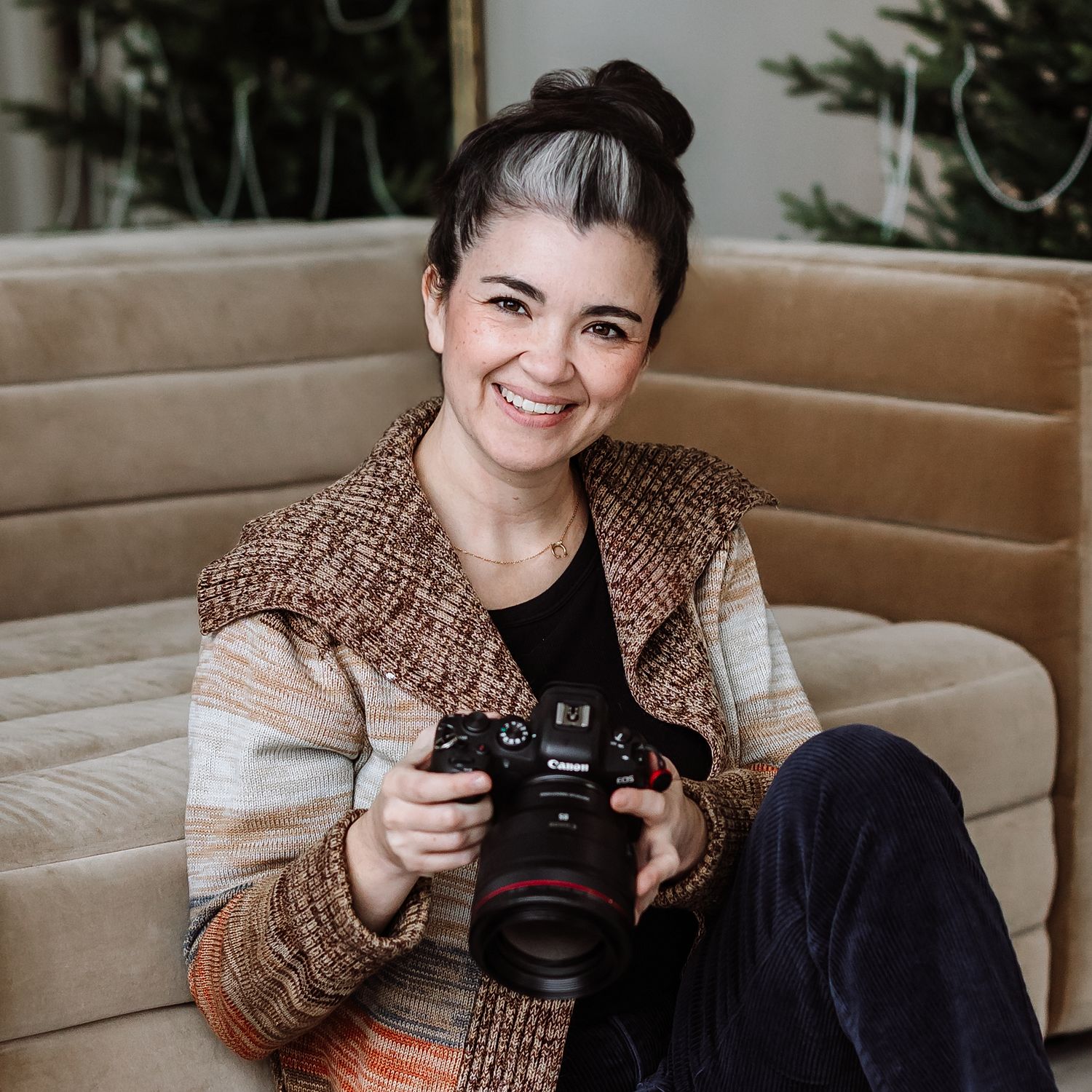 Smiling female photographer holding a Canon DSLR camera while sitting on a beige sofa in a cozy indoor setting.