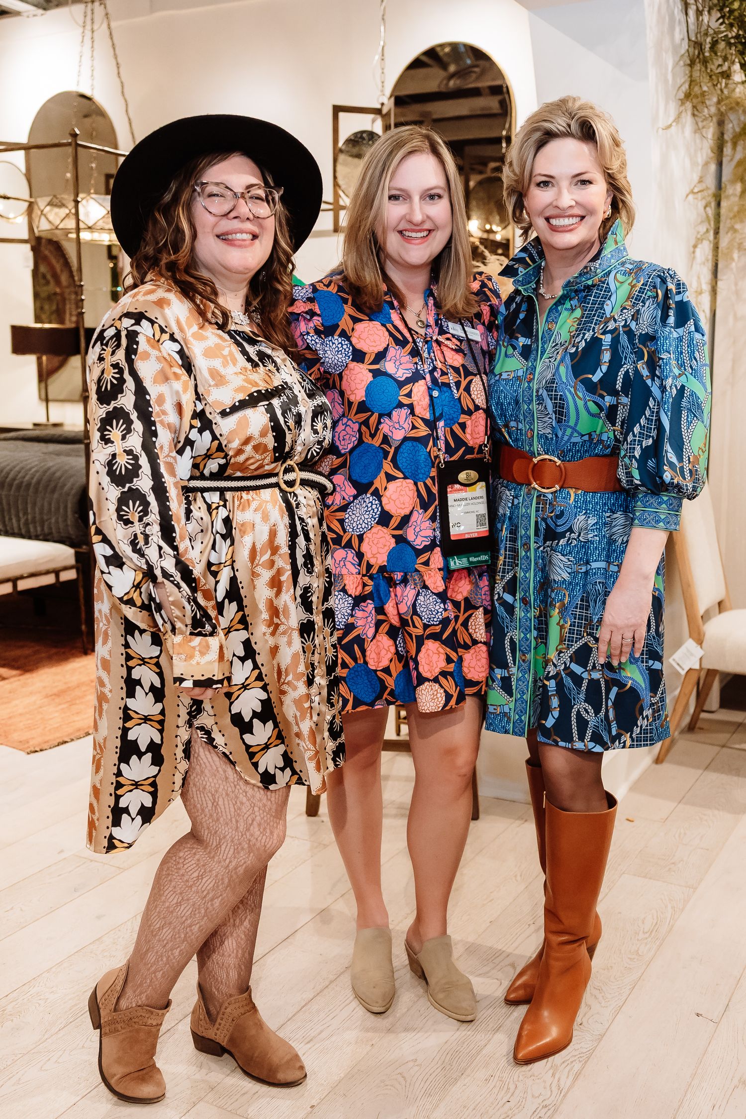 Three women in colorful patterned dresses pose together smiling at an interior design trade event.