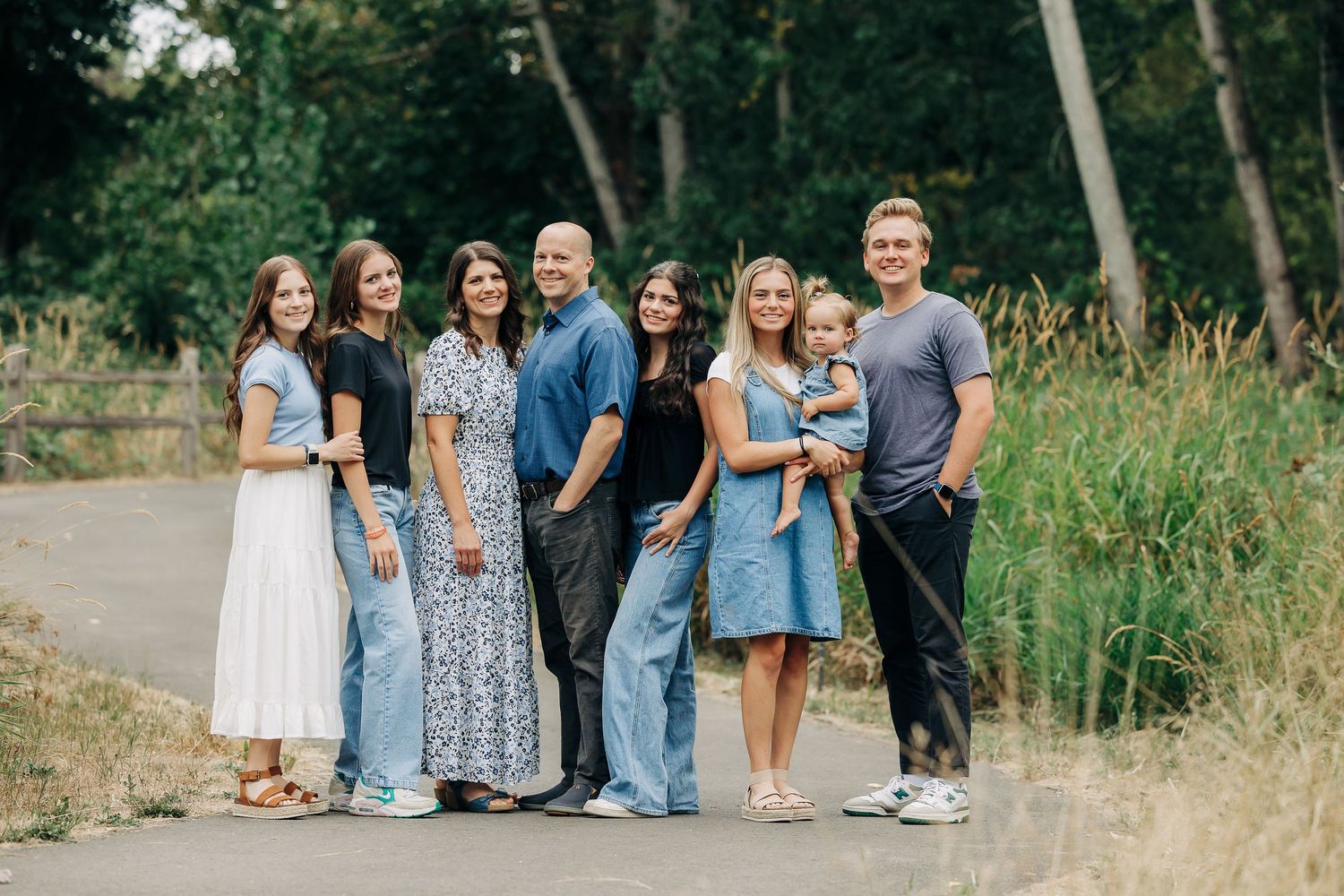 A large family of seven poses together on a path surrounded by lush green trees for an outdoor portrait.