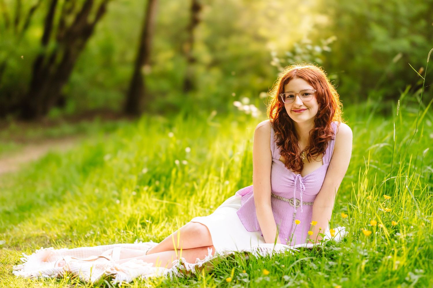 Young redhead woman in lavender dress sitting on blanket in sunny green park, smiling outdoors.