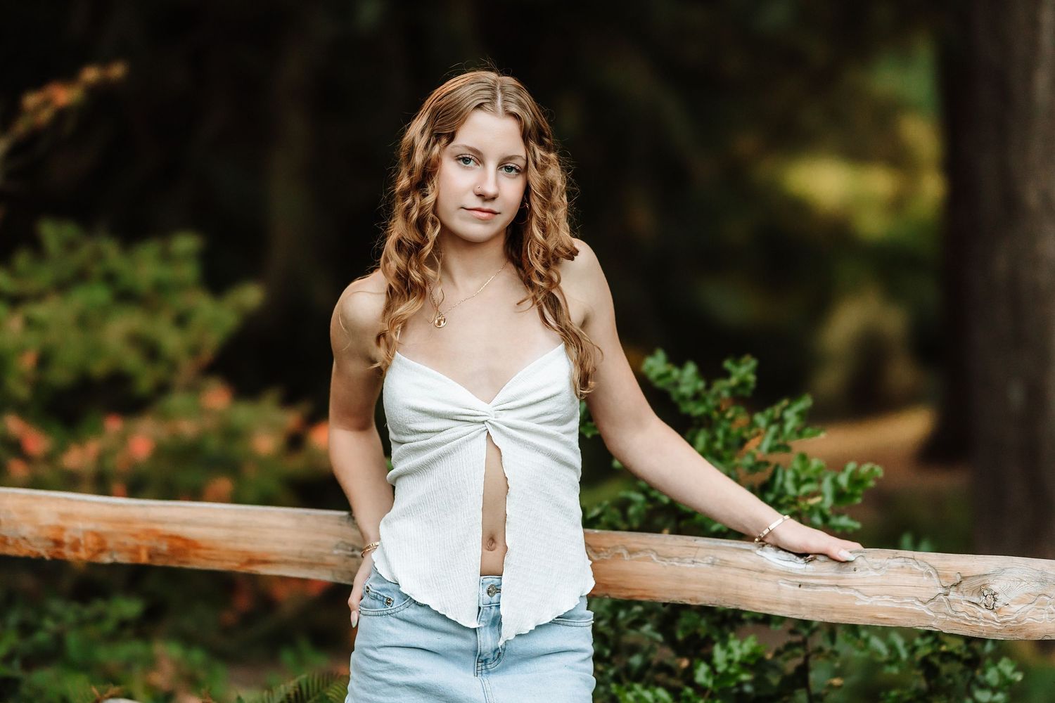 Young woman in white crop top and denim skirt poses by wooden fence in lush forest setting.