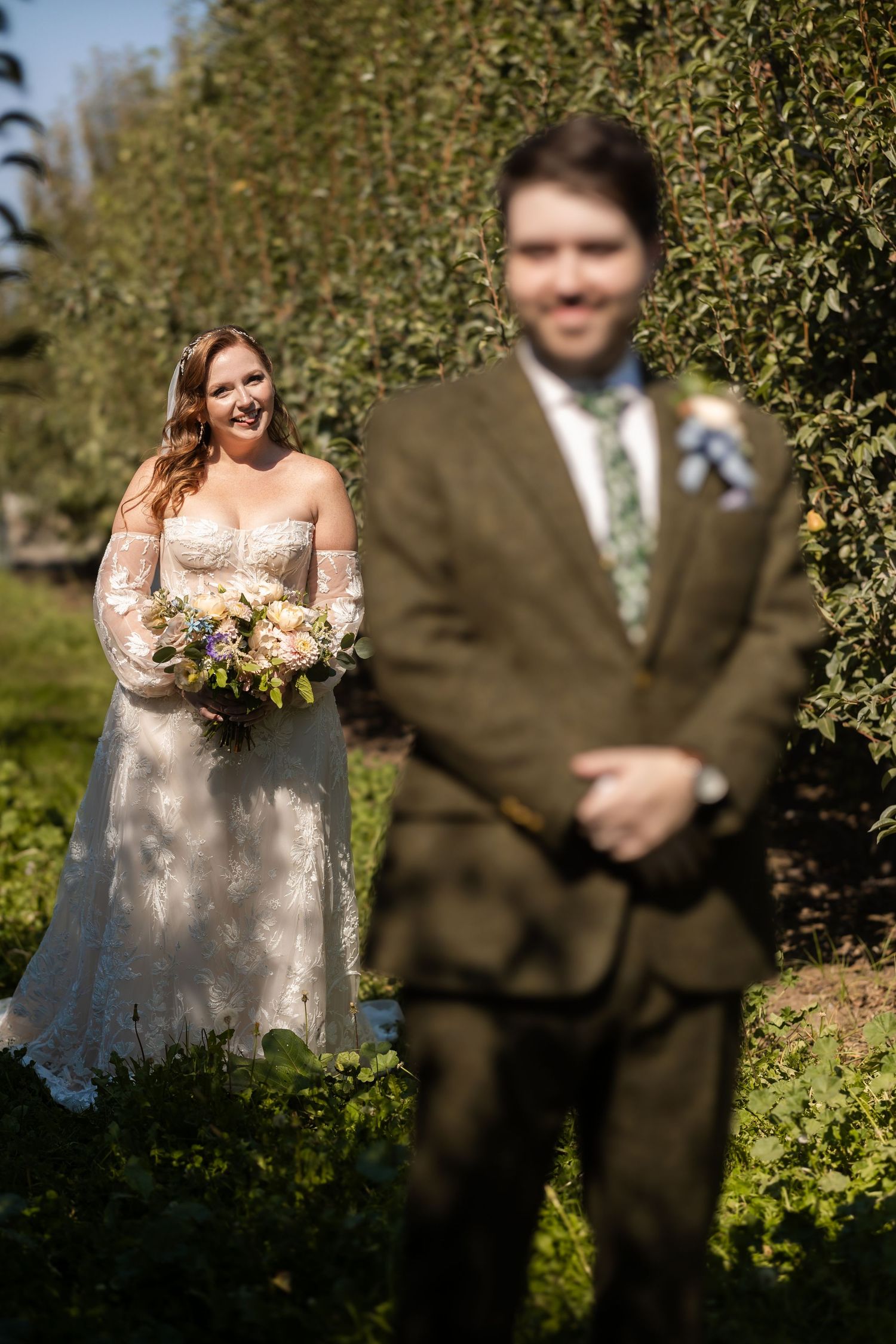Bride in lace gown holding bouquet smiles at blurred groom in green orchard setting.