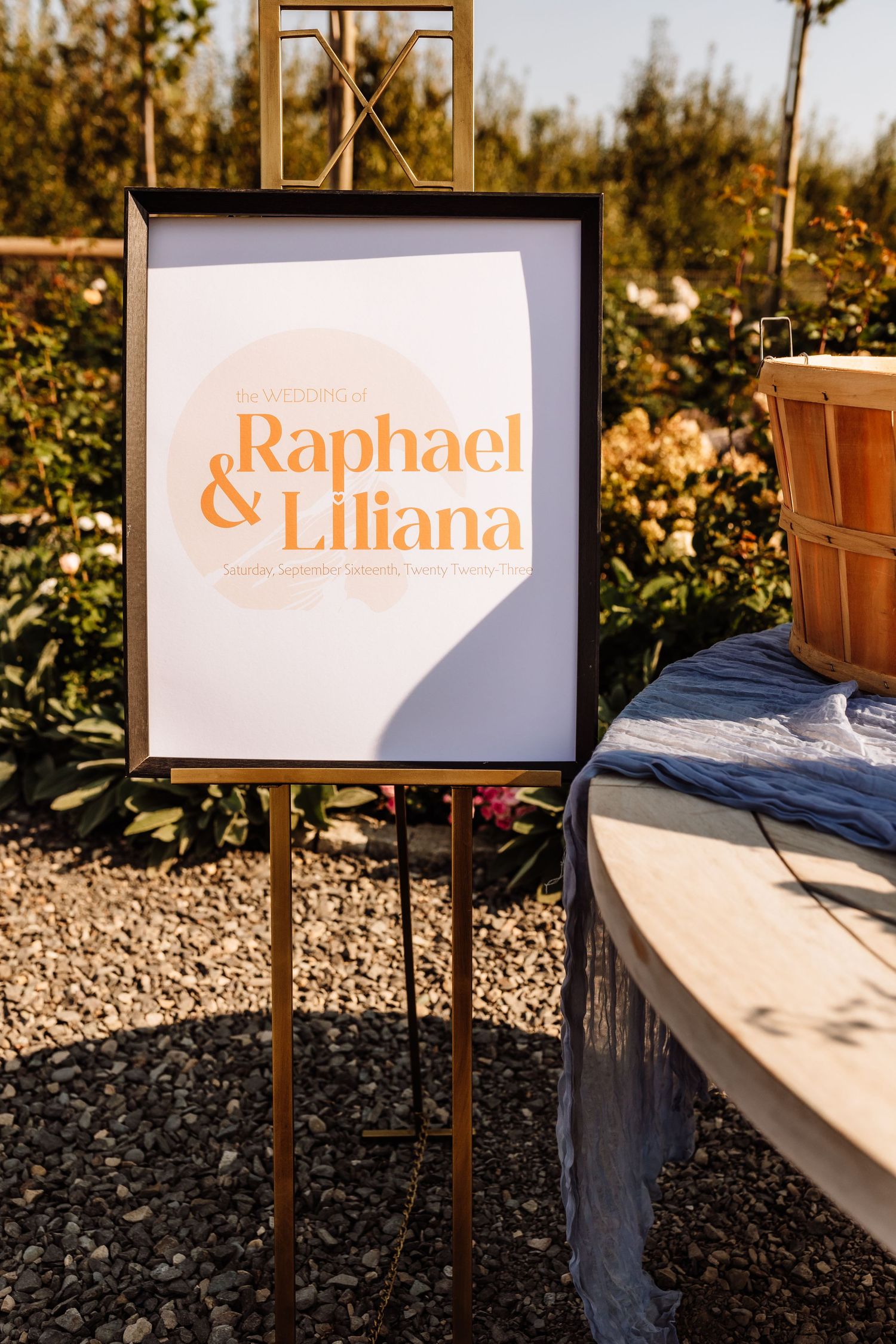 Gold easel displaying modern orange wedding welcome sign for Raphael and Liliana outdoors in garden setting.
