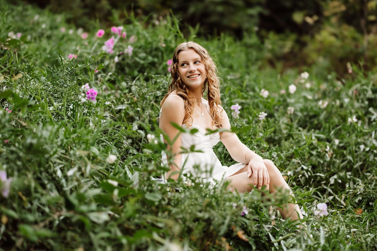 Young woman with curly hair smiling in a lush green meadow with pink wildflowers on a sunny day.