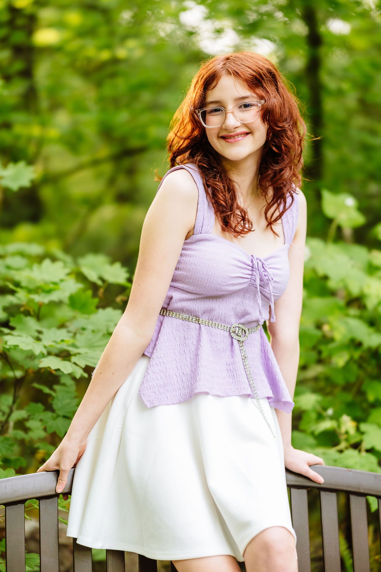 Young woman with red hair in purple top and white skirt smiling outdoors in a lush green forest setting.