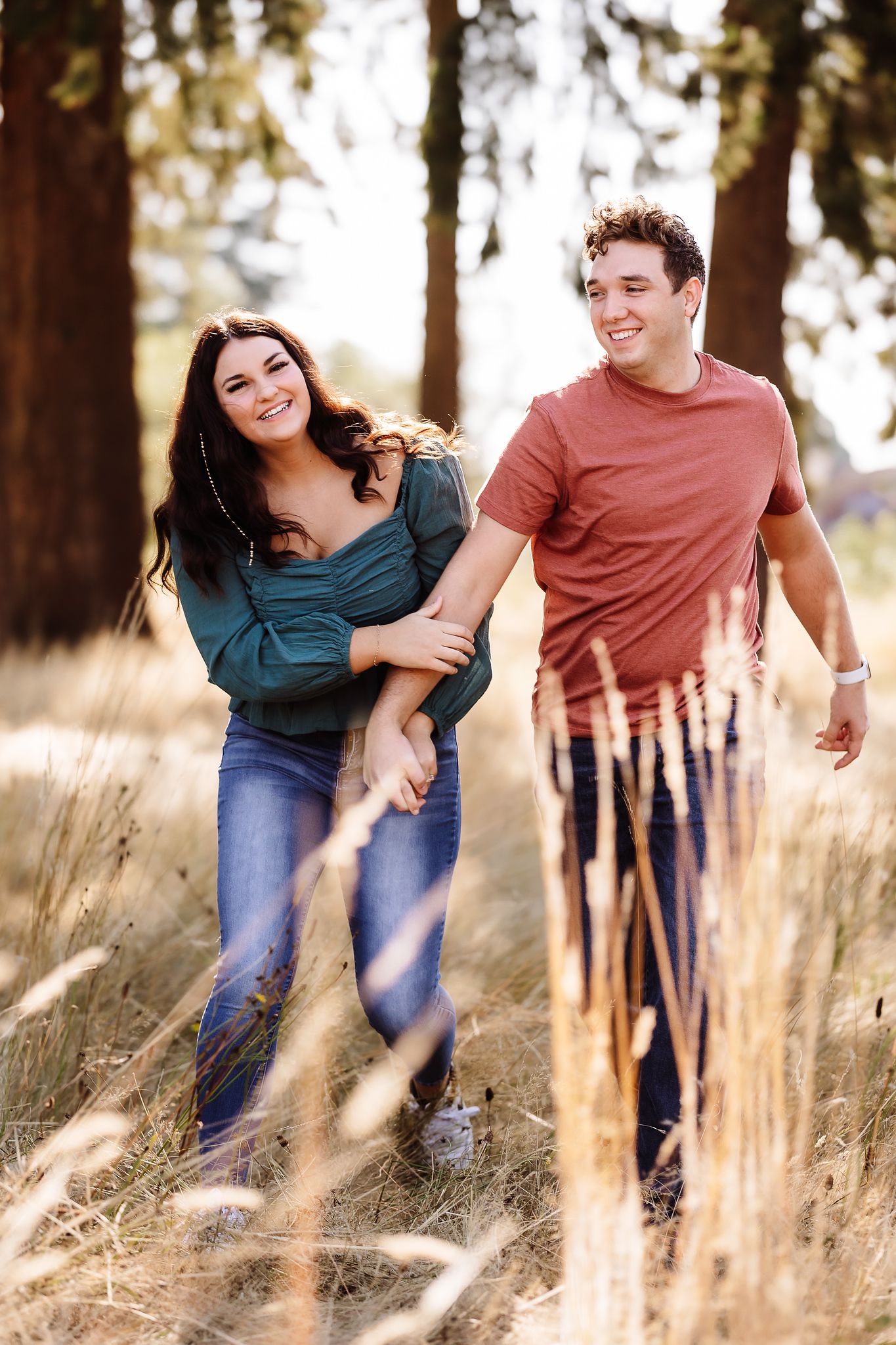 Happy couple walking hand in hand through a sunlit field with tall grass and pine trees in the background.