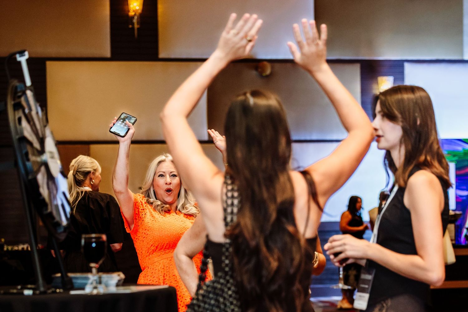 Excited woman in orange dress raises hands at lively indoor networking event with guests.