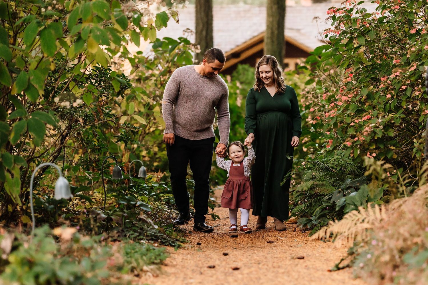 Family of three walking on a garden path, pregnant mother in green dress, father in gray sweater, toddler between them.