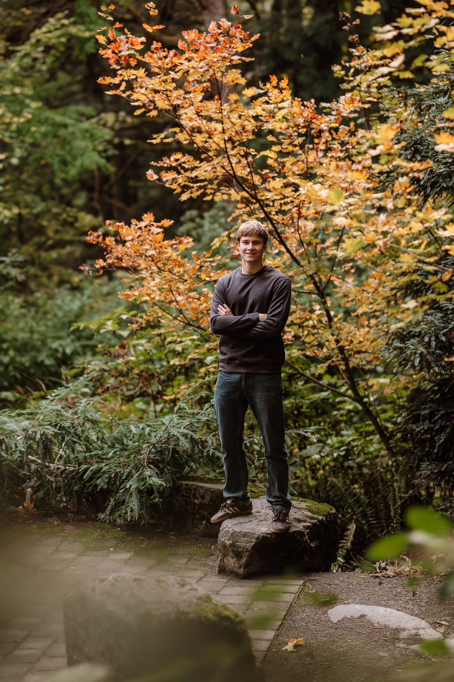 Young man standing on a rock with arms crossed, surrounded by autumn foliage in a lush garden setting.