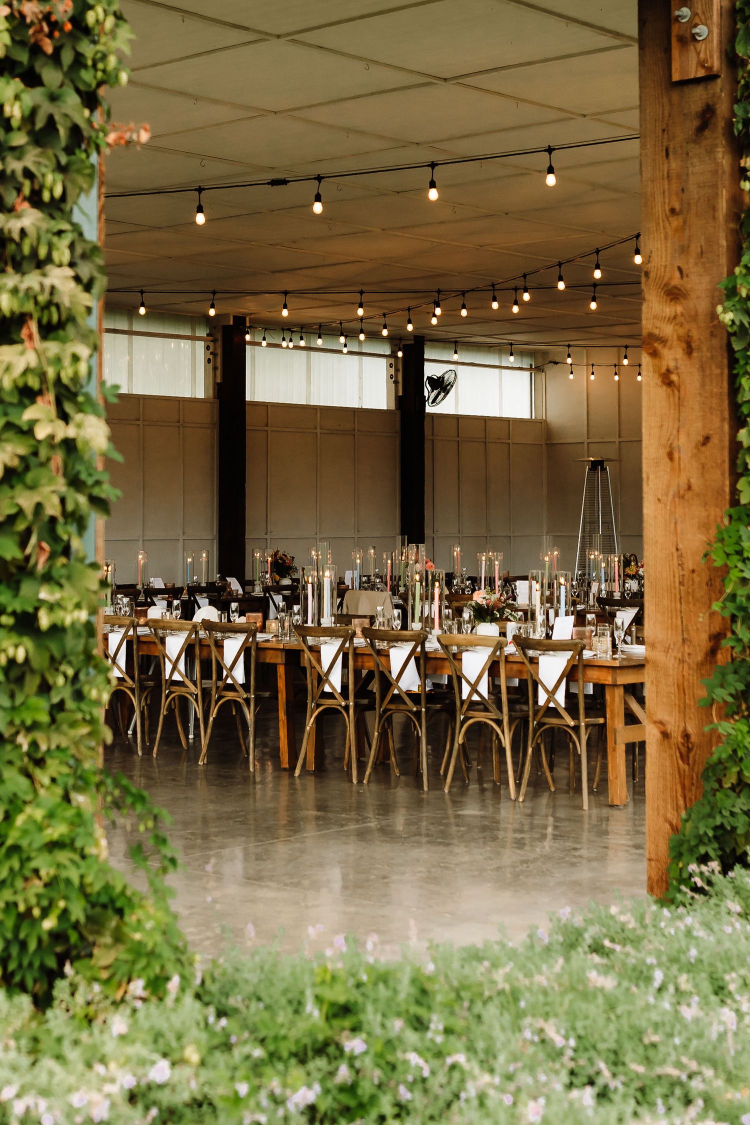 Elegant wedding reception hall with string lights, long farm tables, cross-back chairs, and lush greenery framing the entrance.