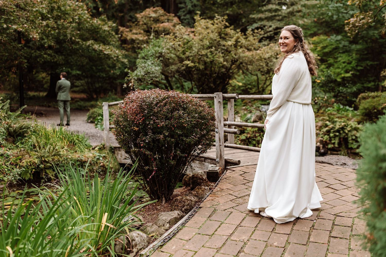 Bride in white gown smiles over shoulder on garden path with wooden bridge and lush greenery.