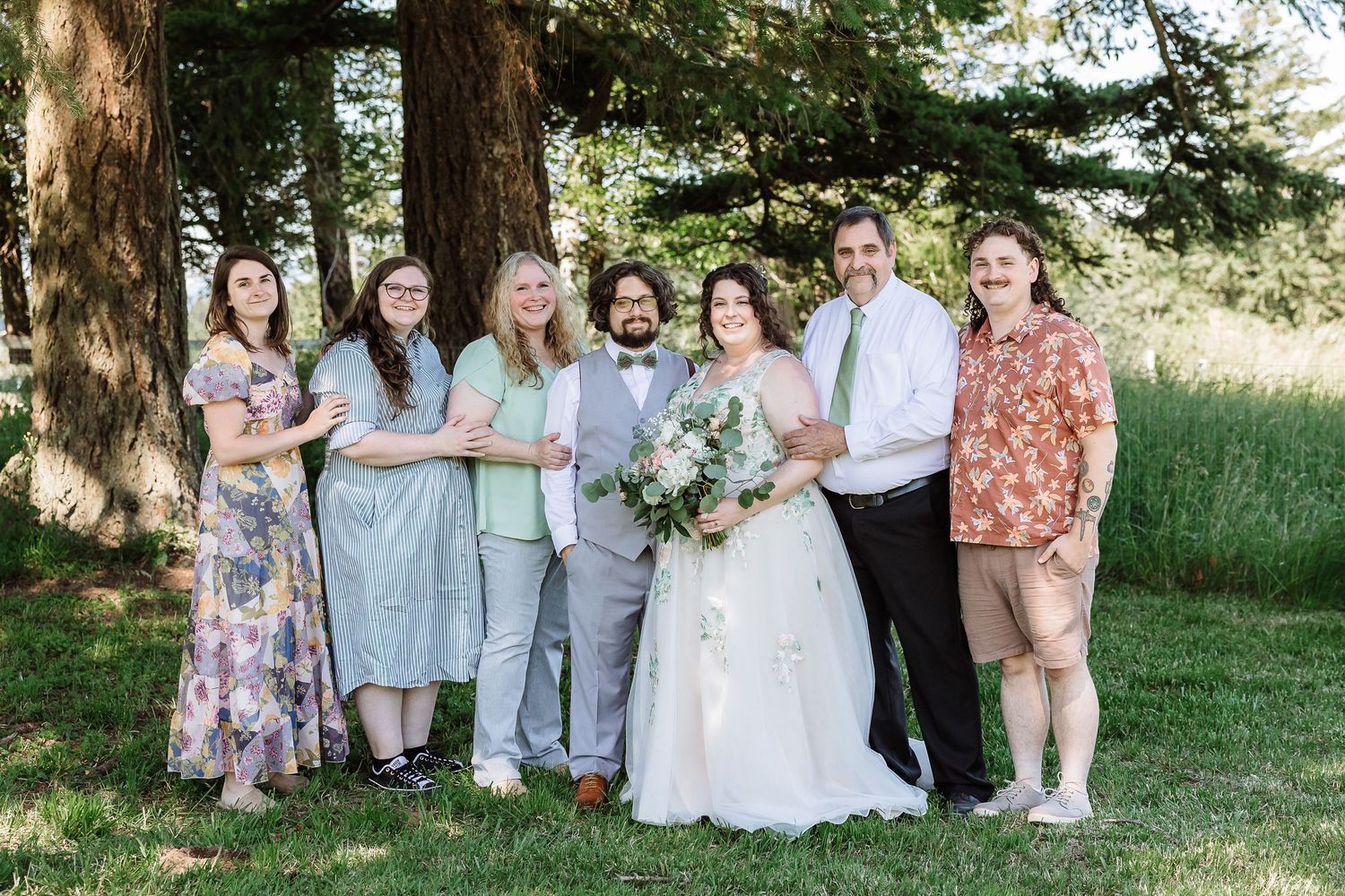 Wedding party poses under tall trees in a lush green park, bride holding a white bouquet.