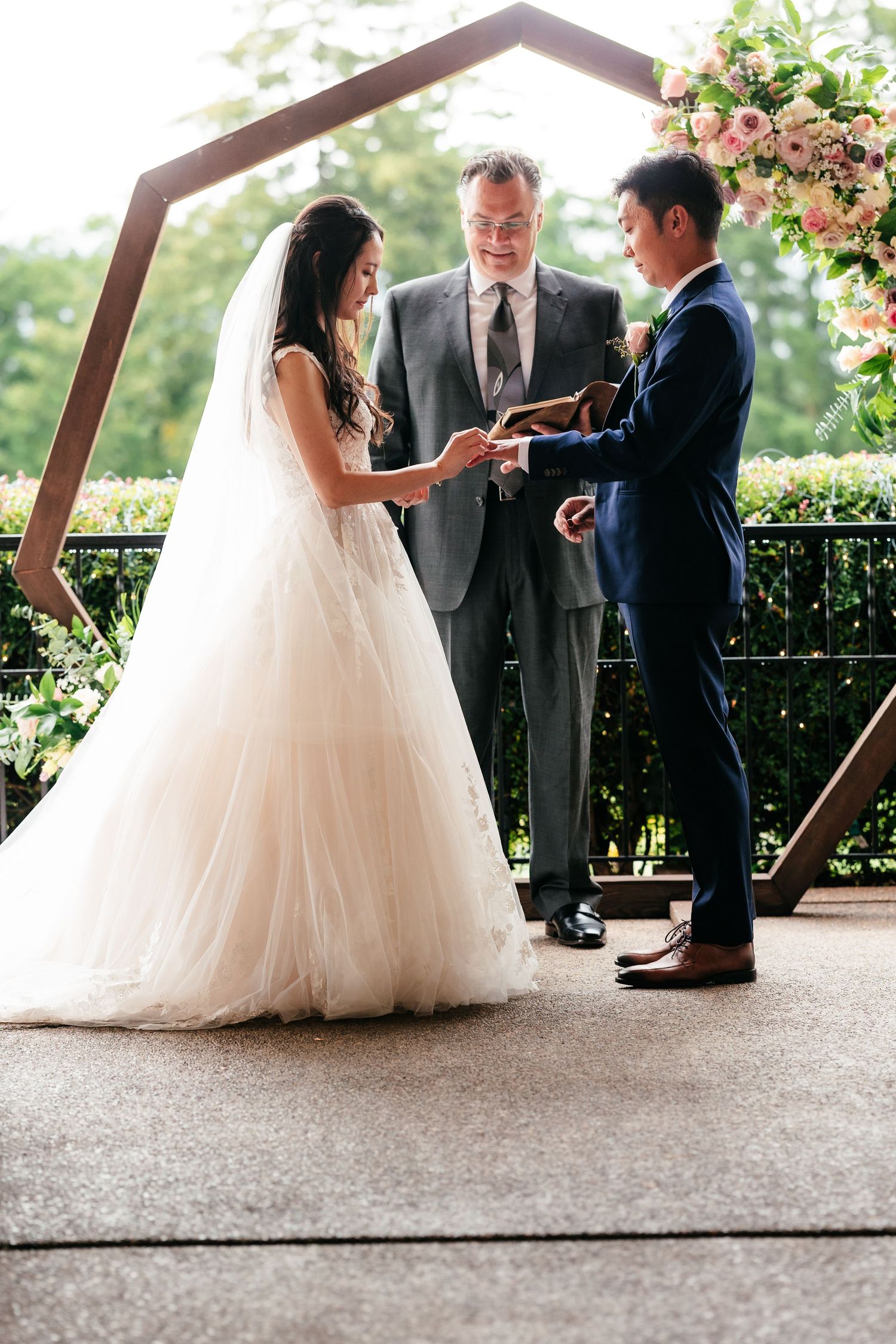Bride places ring on grooms finger before hexagonal wooden arch at outdoor wedding ceremony.
