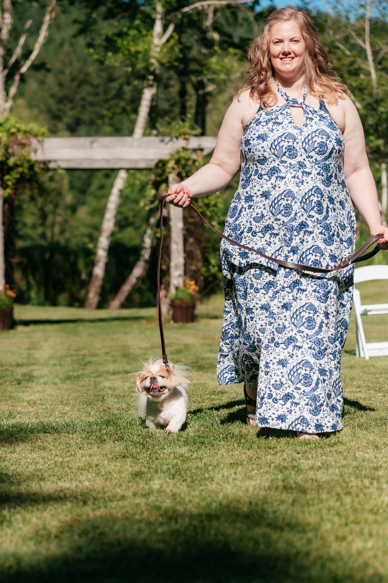 Woman in blue floral dress walks small fluffy dog on leash at outdoor wedding venue.