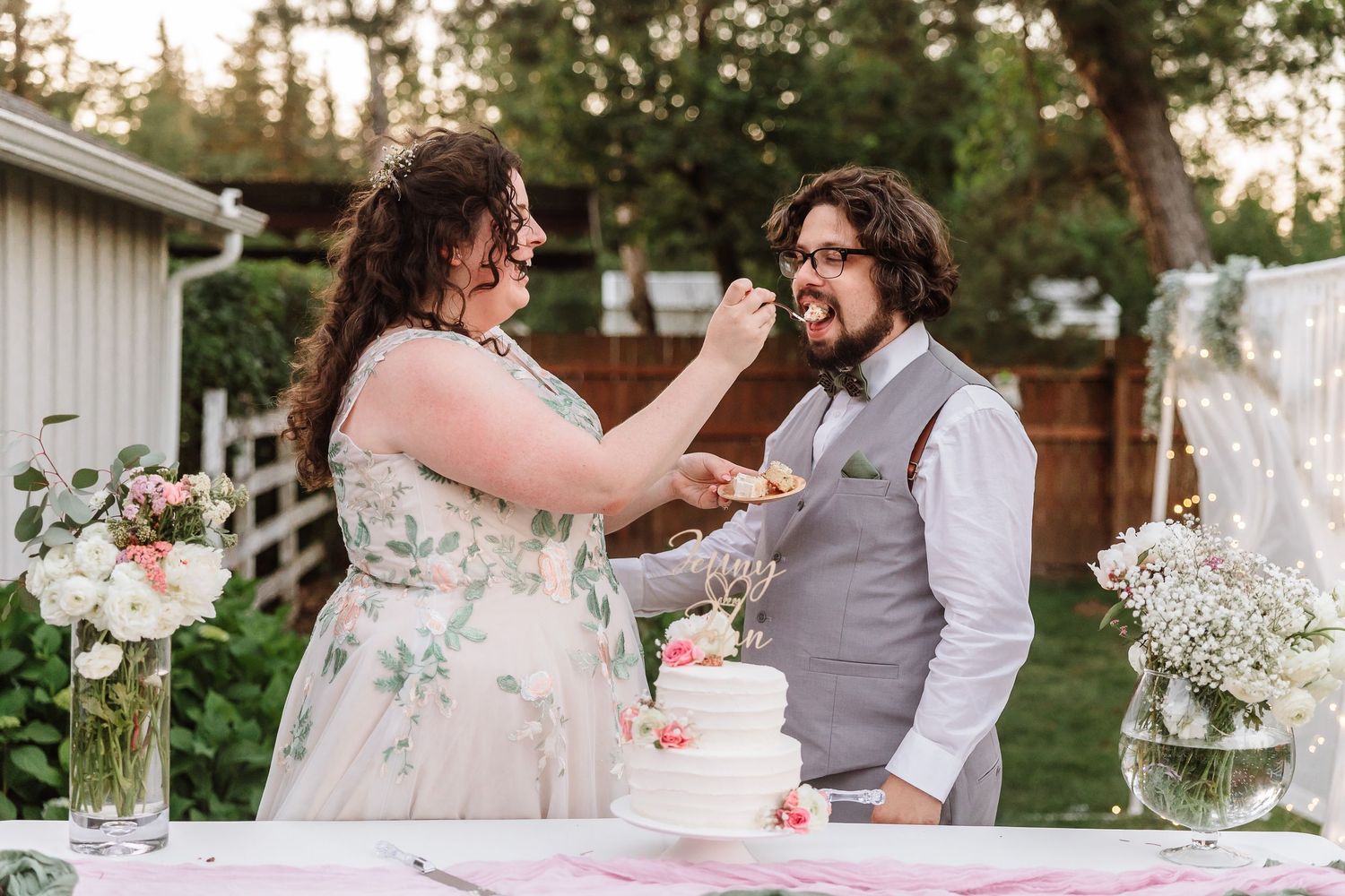Bride feeds groom wedding cake at outdoor backyard reception with floral arrangements on table.