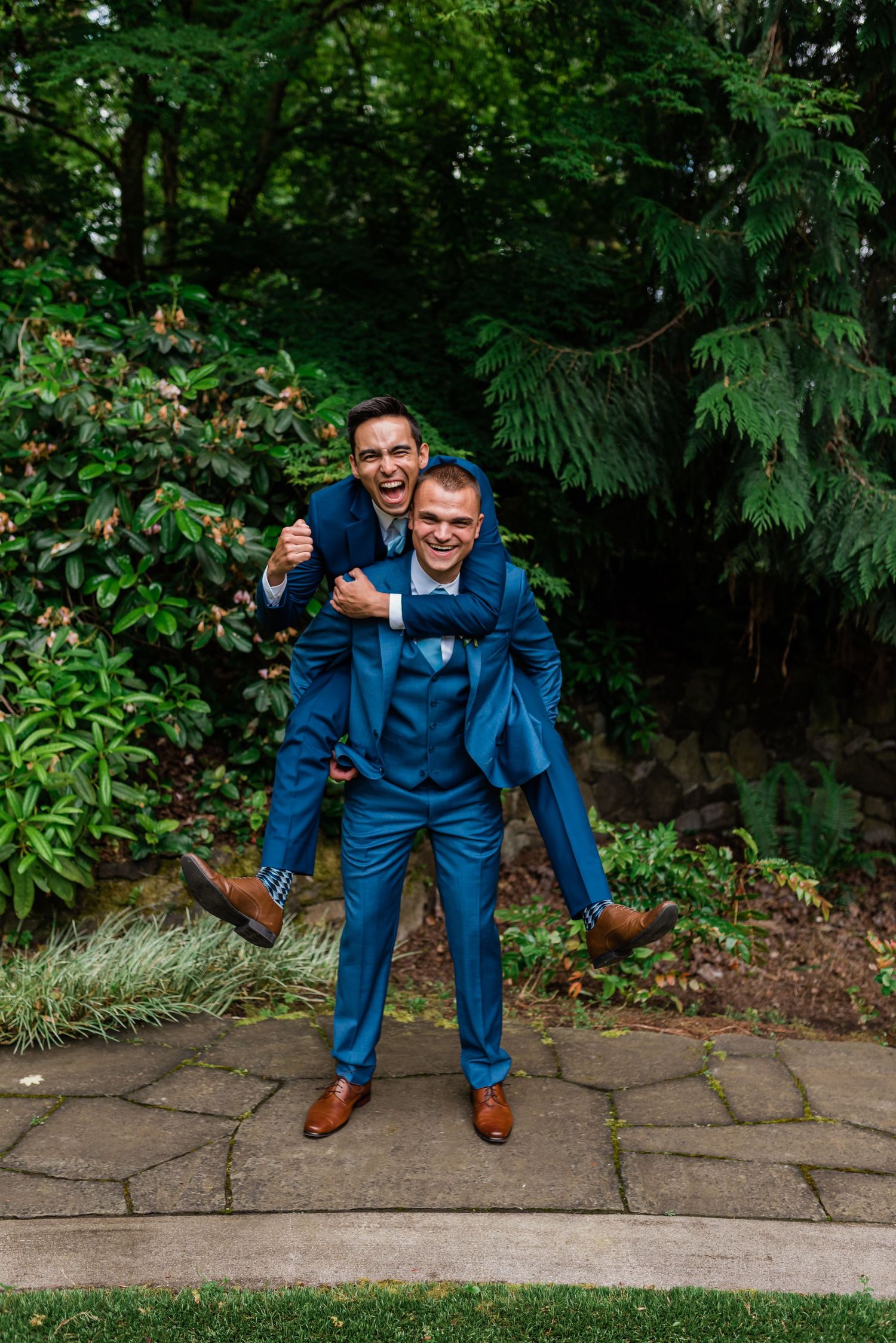 Two groomsmen in navy blue suits laughing as one gives the other a piggyback ride outdoors.