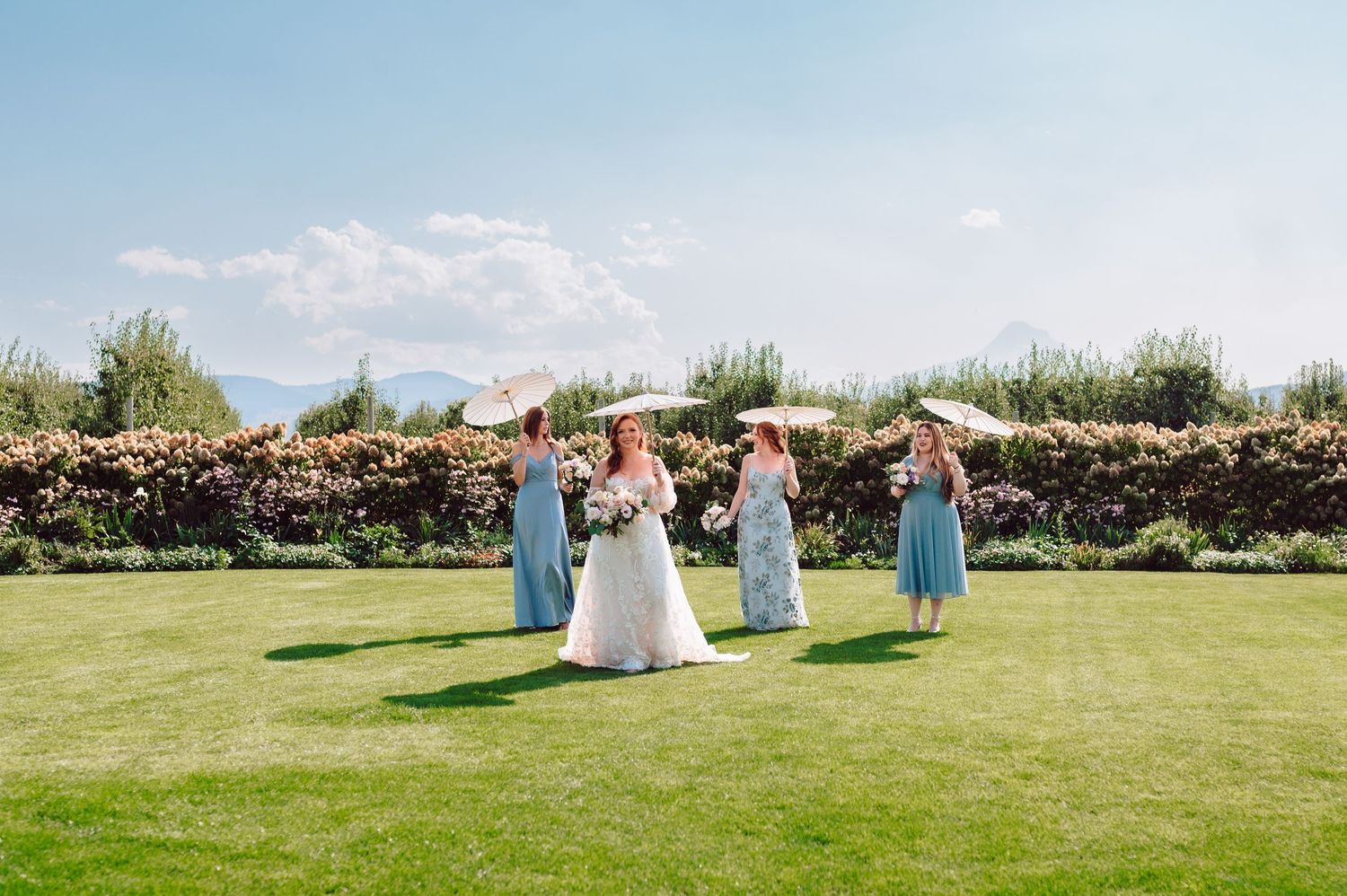 Bride in white gown poses with bridesmaids in blue dresses holding parasols on a sunny lawn.