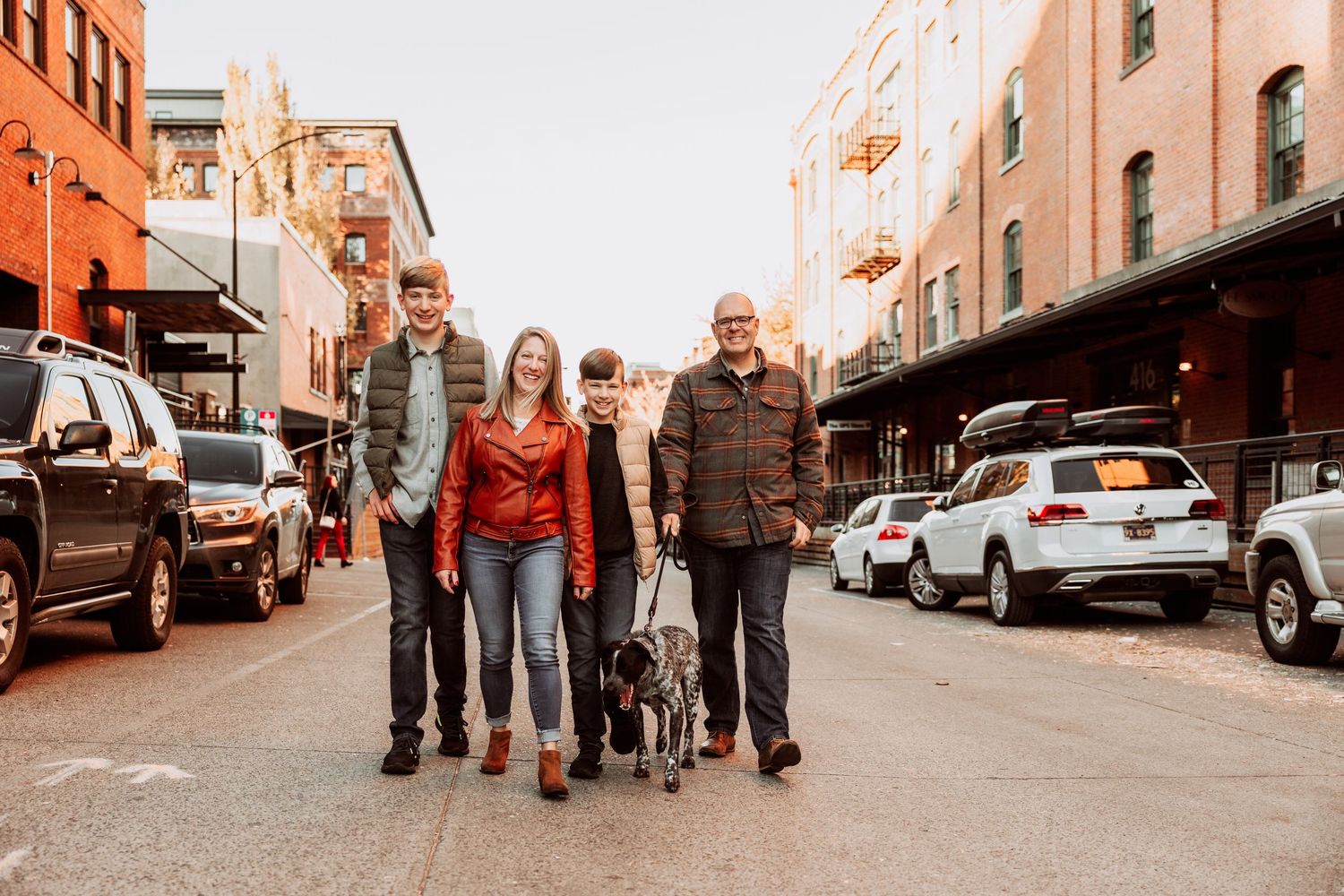 Family of four with a dog walking on a city street at sunset, surrounded by parked cars and urban buildings.