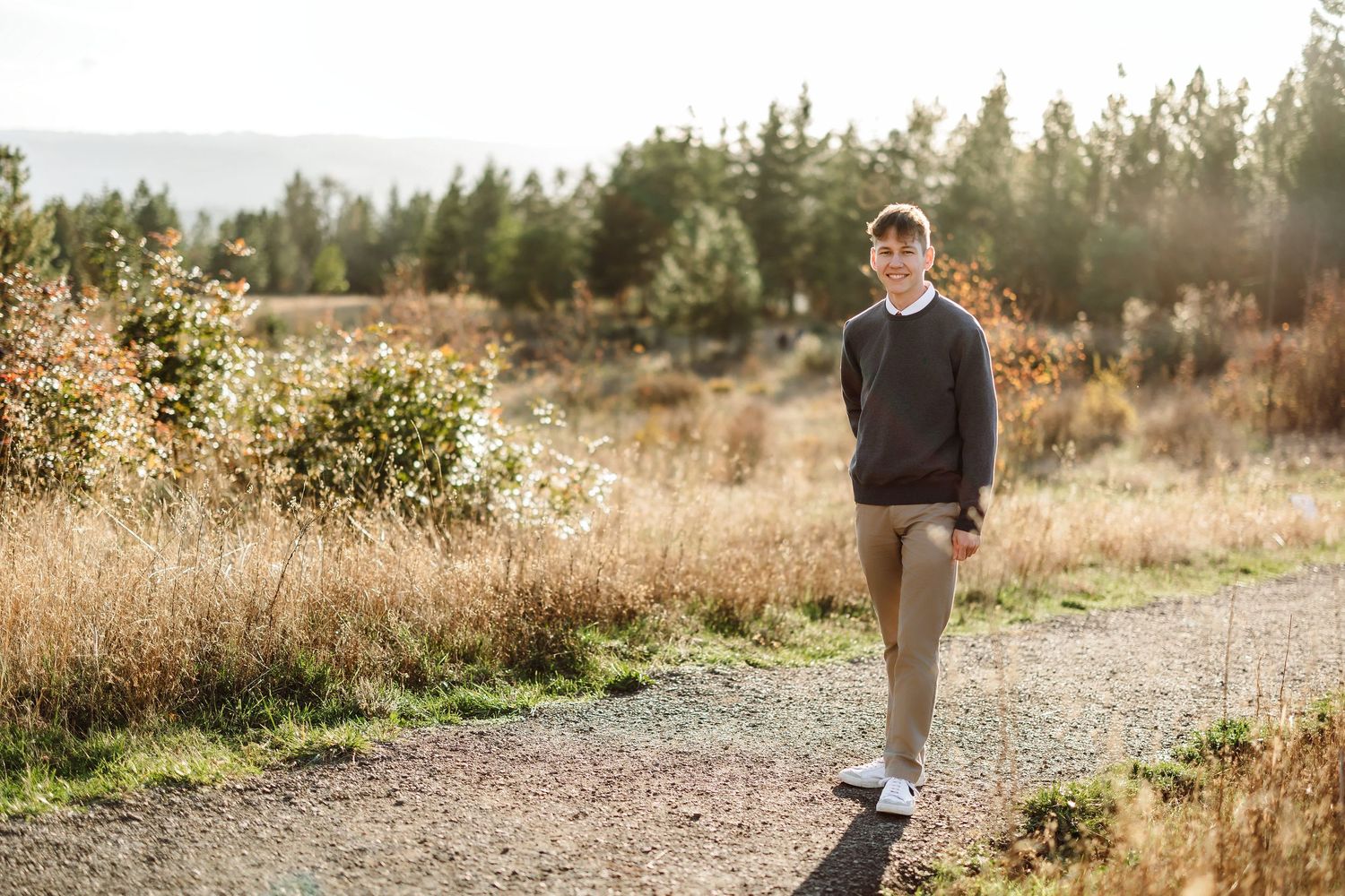 Young man in gray sweater and khaki pants walking on a dirt path surrounded by autumn foliage and pine trees.