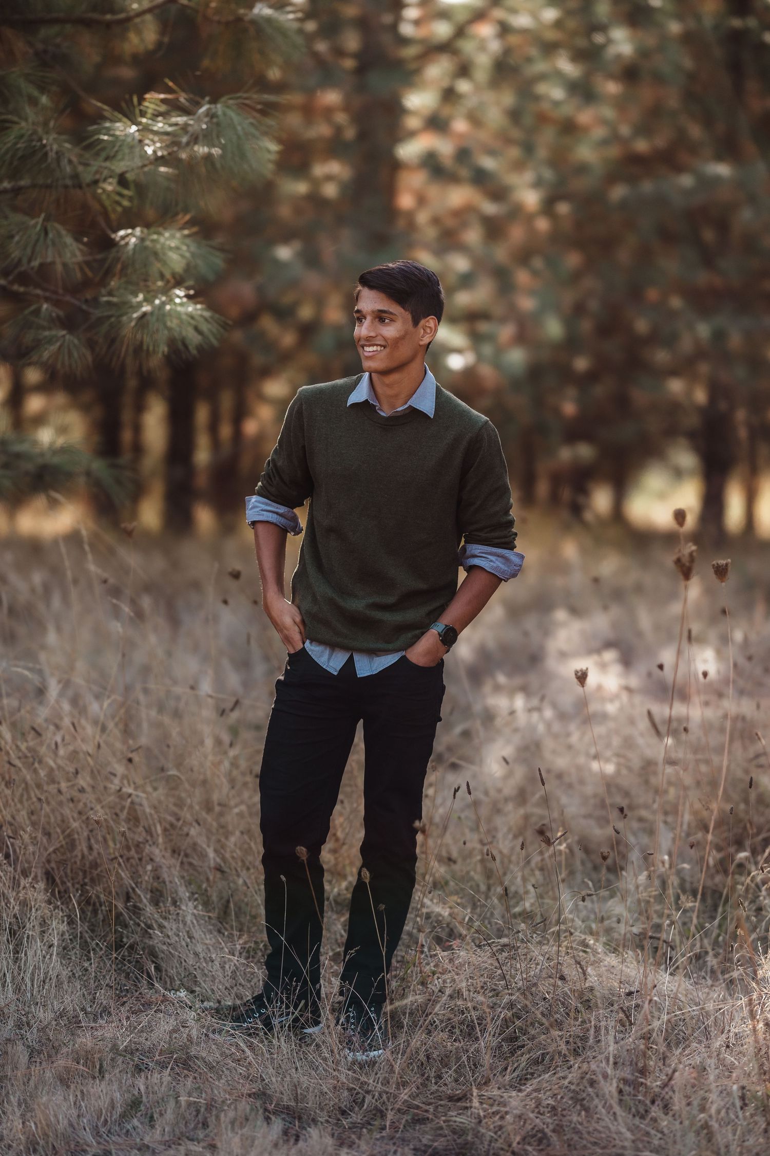 Young man in olive sweater and black jeans smiling outdoors in a forest clearing with dry grass and pine trees.