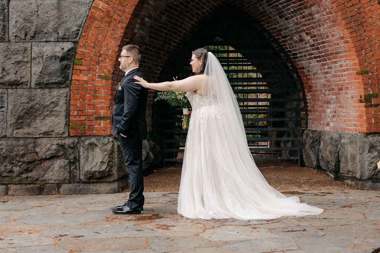 Bride and groom share a tender first look moment under a rustic brick archway on stone pavement.
