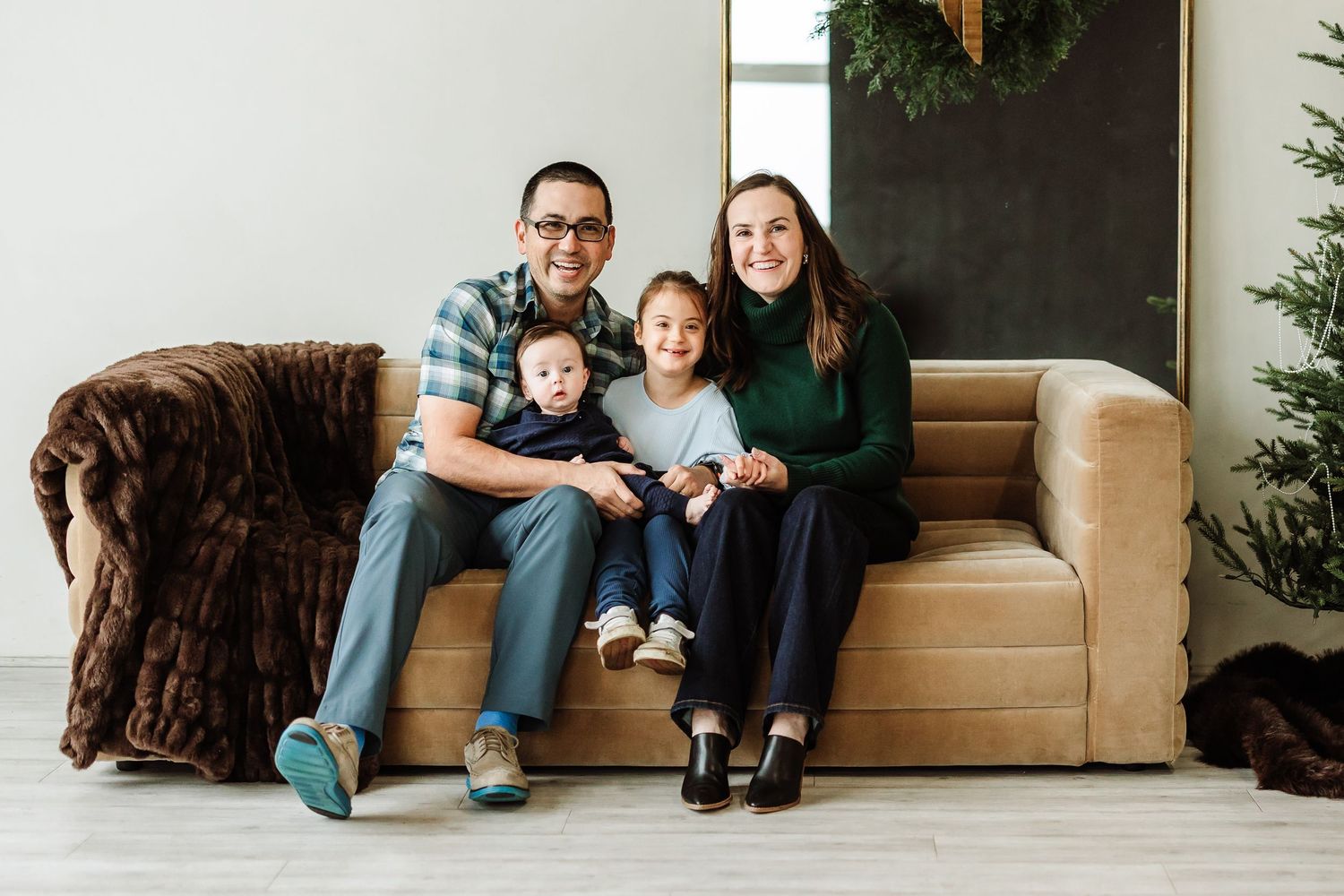 A happy family of four poses on a tan sofa with Christmas decorations in a bright white studio setting.
