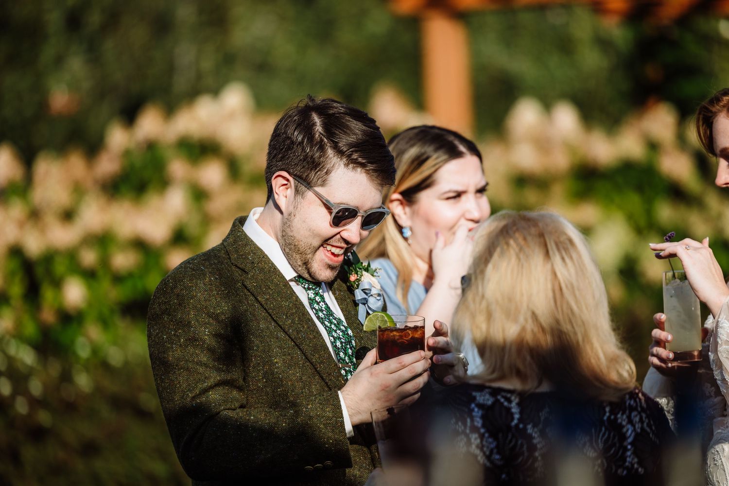 A man in a green tweed suit and sunglasses smiles while holding a drink at an outdoor wedding reception.