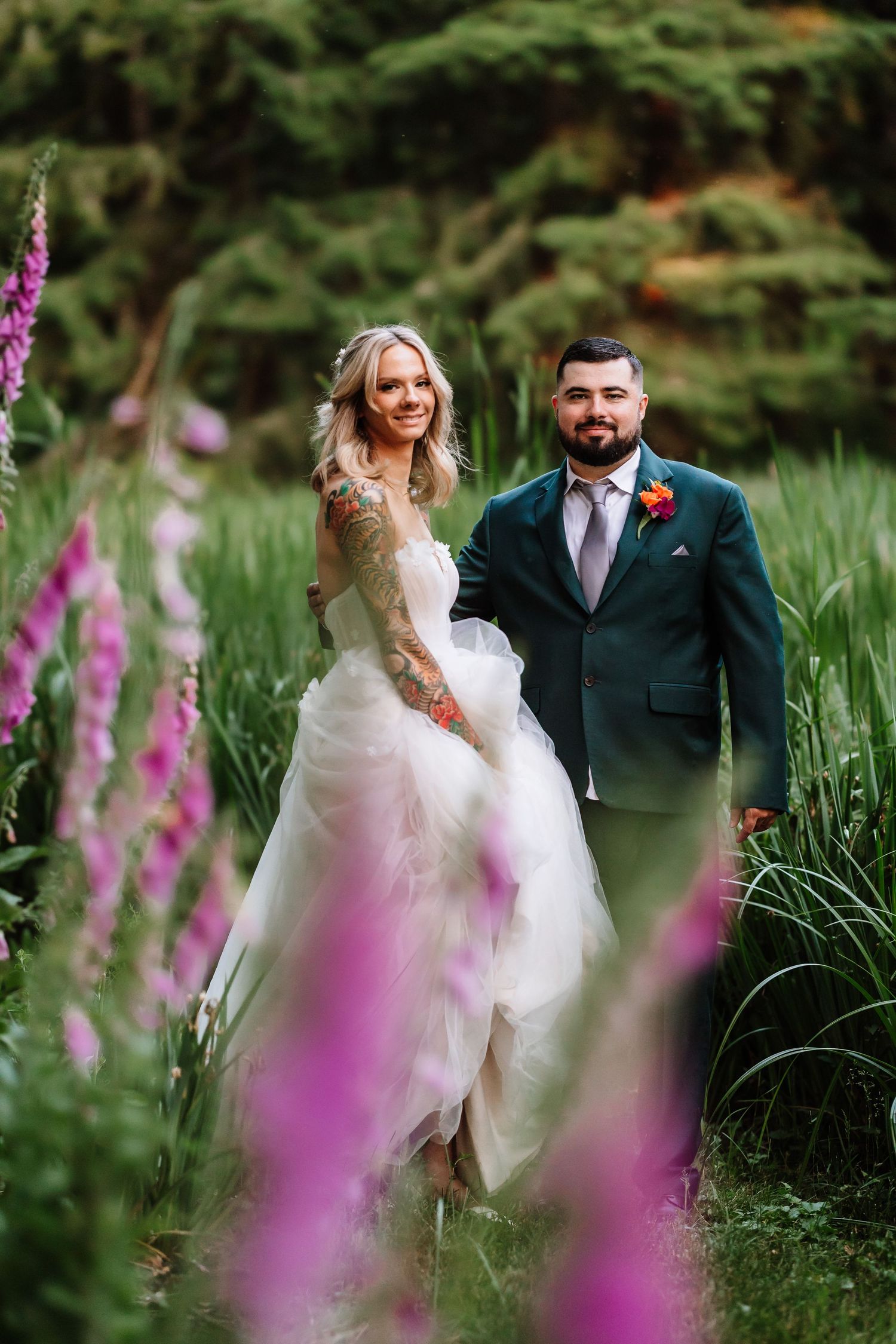 Tattooed bride in white tulle gown walks with bearded groom through vibrant purple foxglove garden.