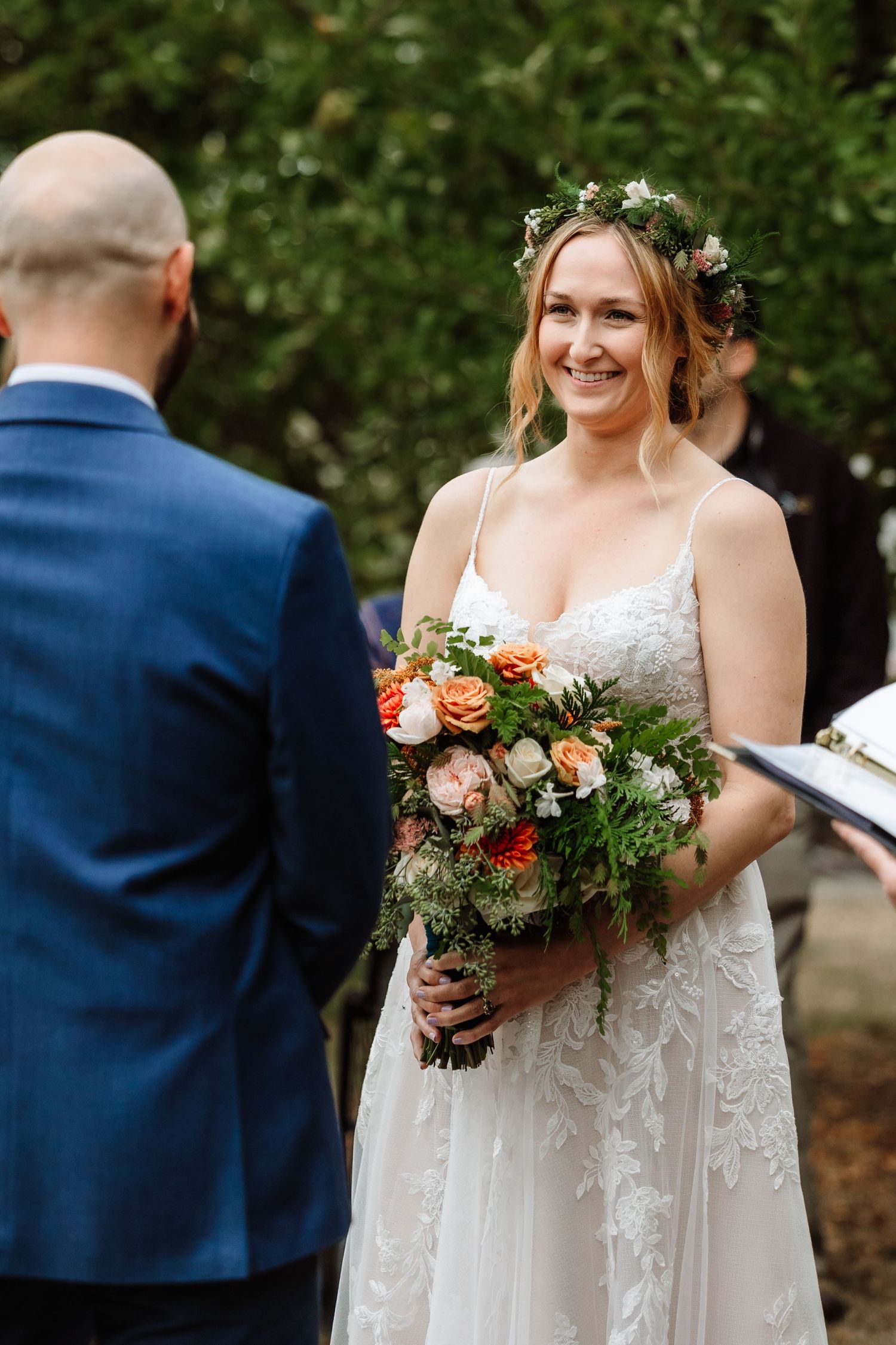 Radiant bride wearing flower crown holds orange and pink bouquet while exchanging vows outdoors.