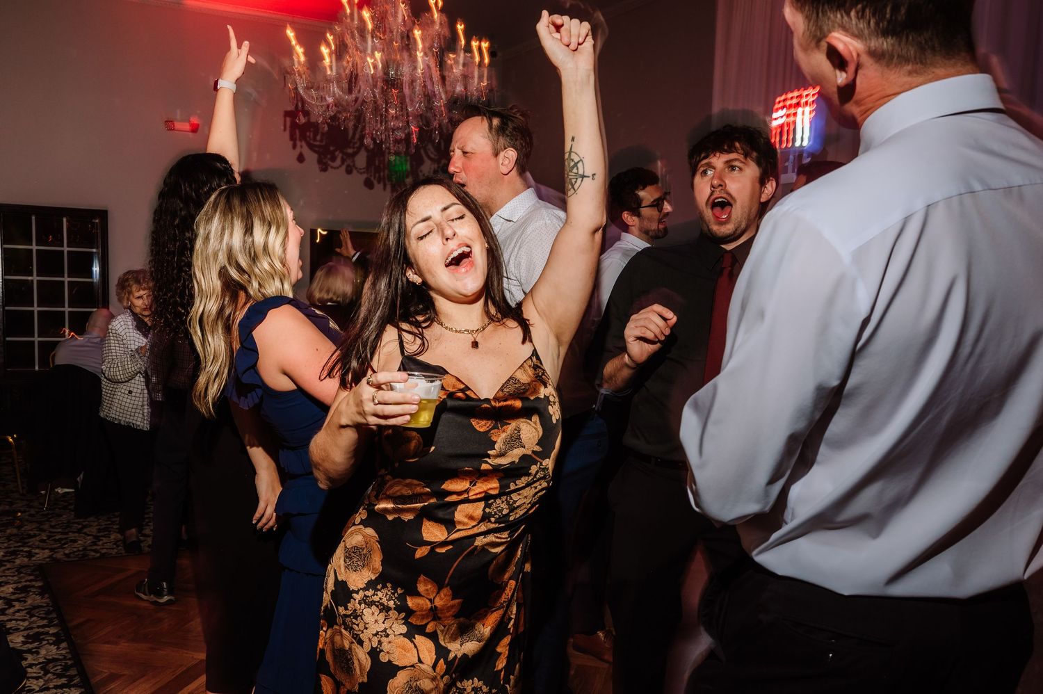 Wedding guest in floral dress dances joyfully with arms raised at lively evening reception party.
