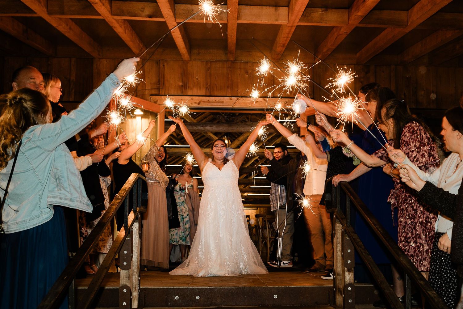 Bride walks through sparkler tunnel exit surrounded by cheering wedding guests in rustic barn venue.