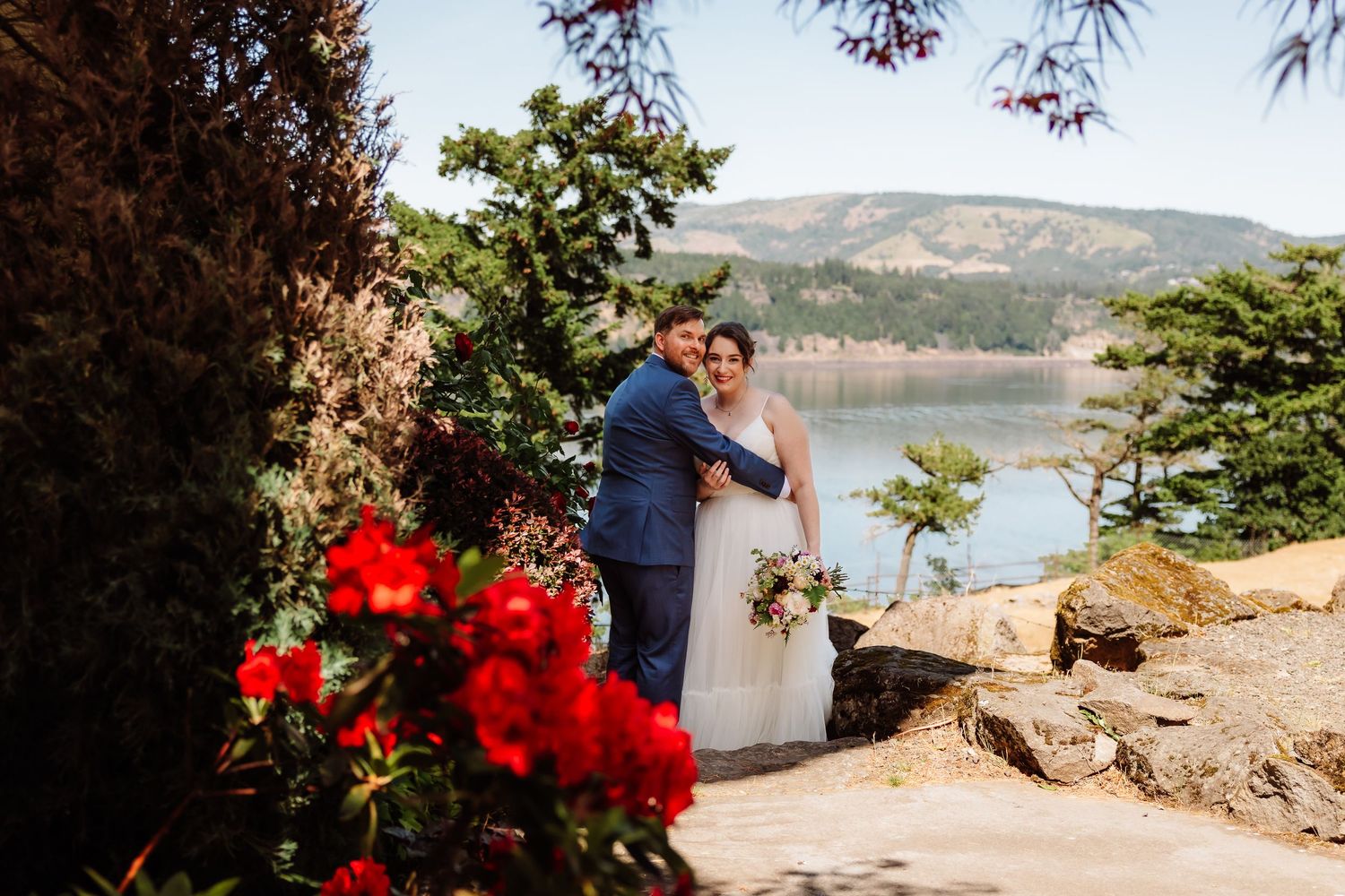 Bride and groom embrace near red flowers with a scenic lake and mountains in the background.