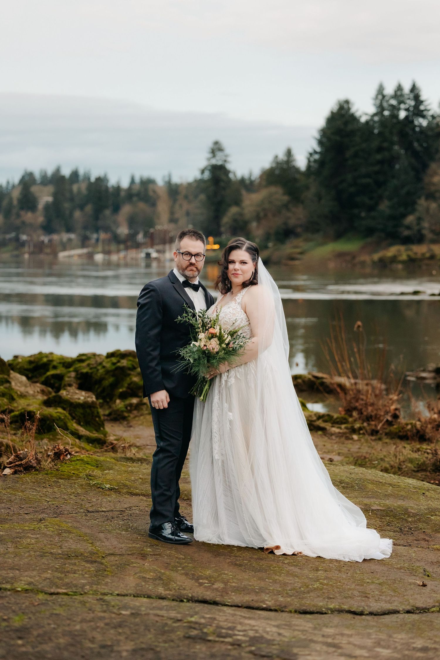 Bride and groom posing by a lake with evergreen trees in an overcast outdoor wedding portrait.