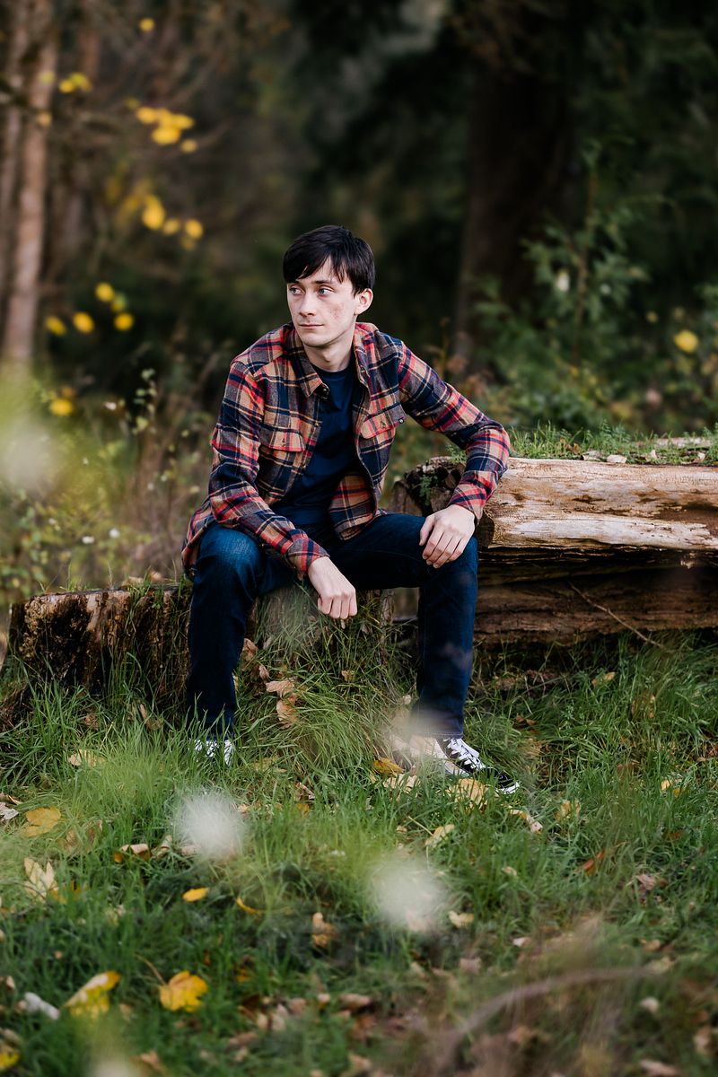Young man in plaid shirt sitting on a fallen log in a lush green forest setting.