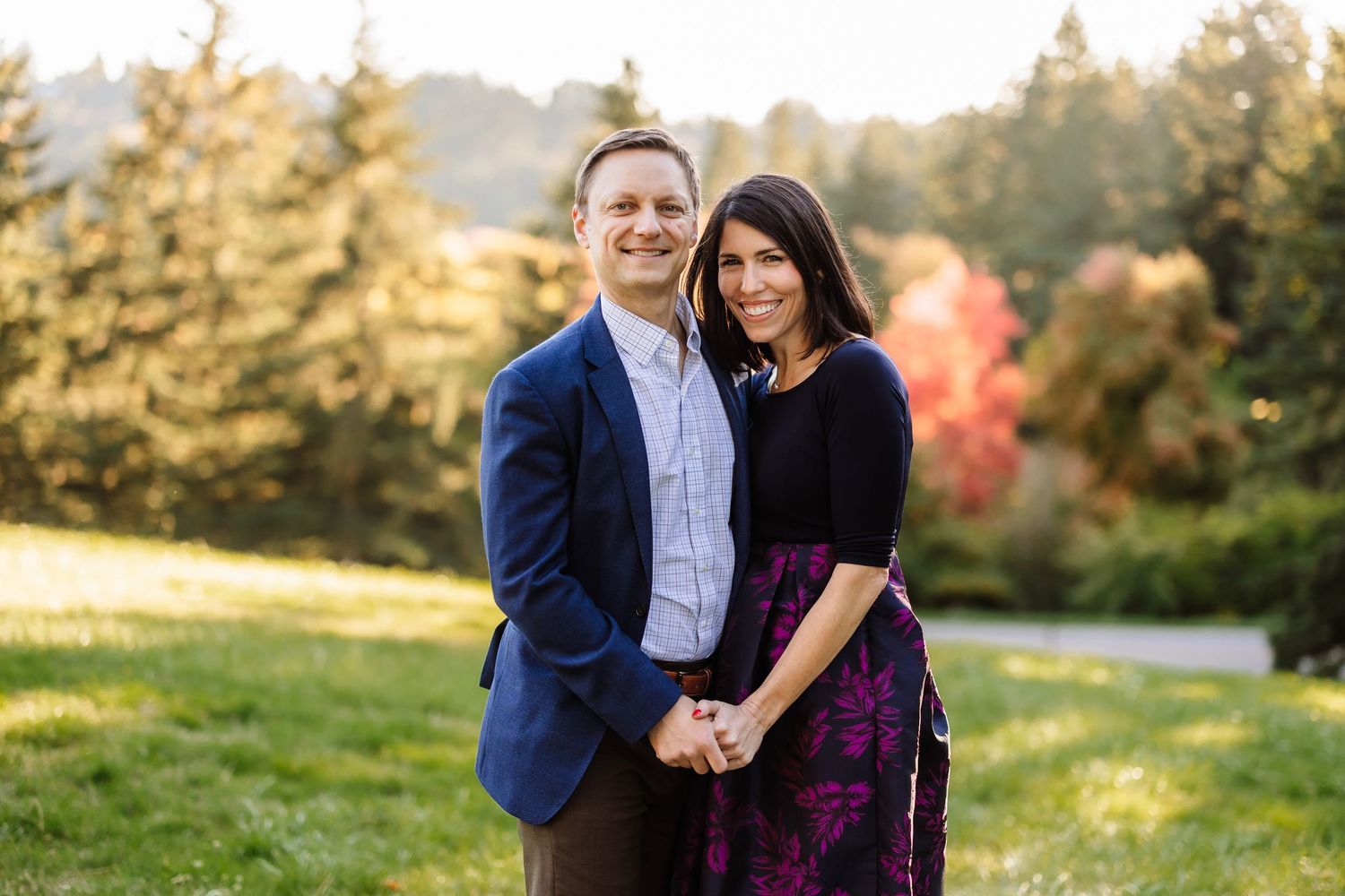 Couple smiling and holding hands outdoors on green grass with colorful autumn trees in golden hour light.
