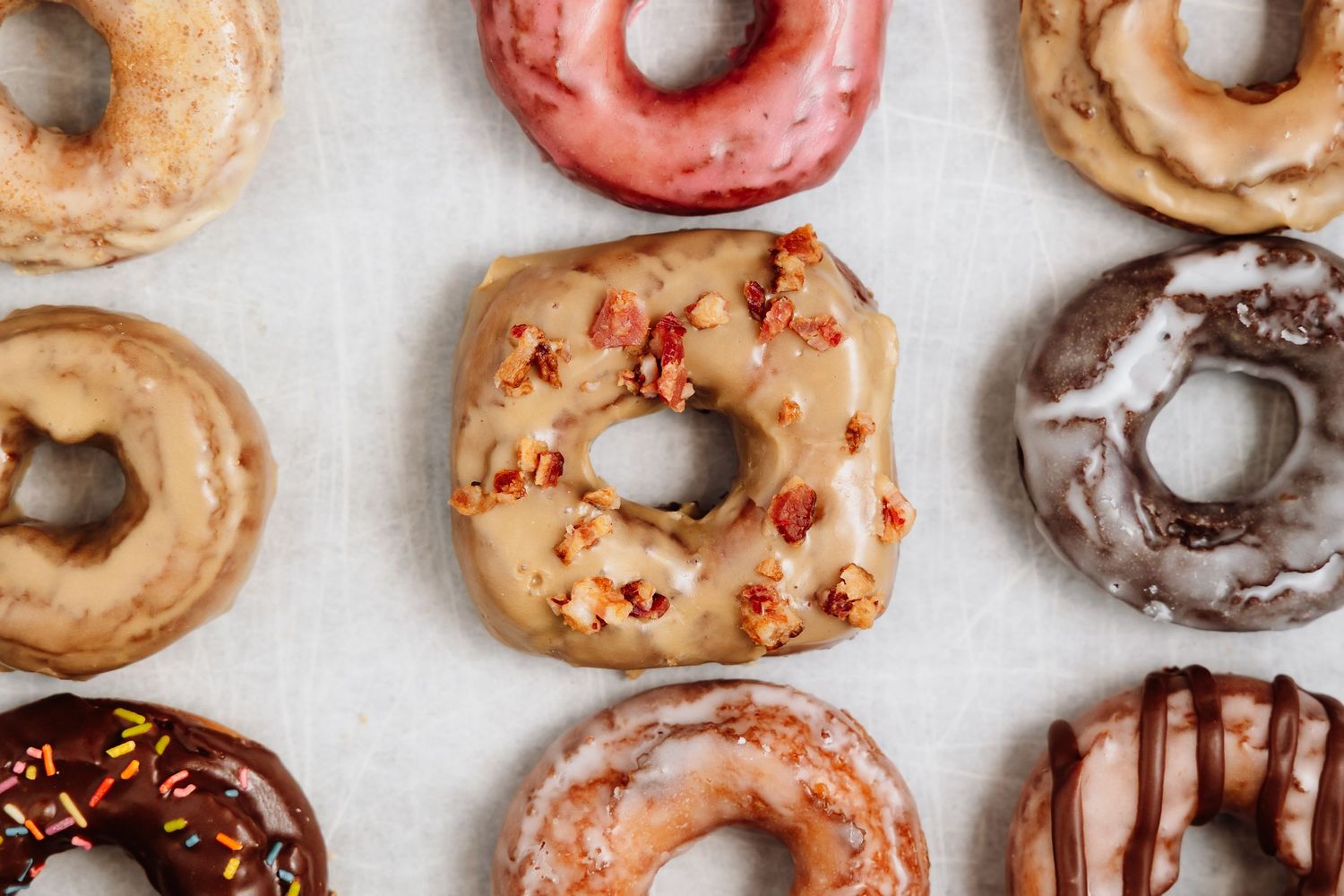 Assorted glazed donuts with chocolate, maple bacon, and strawberry toppings arranged on white surface.