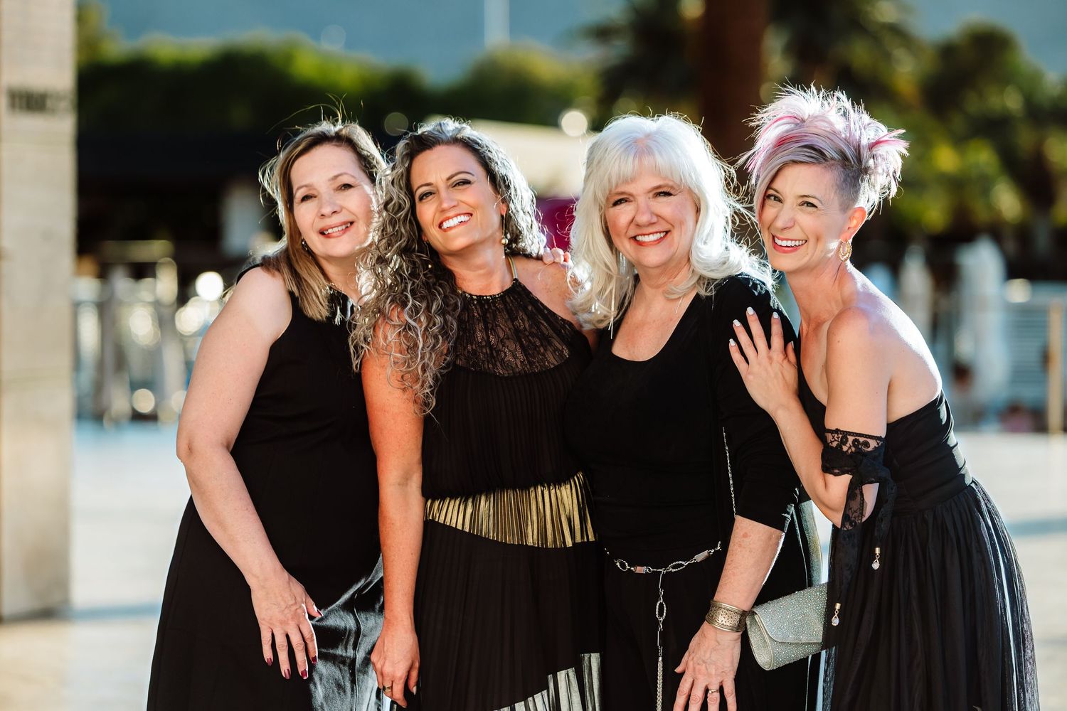Four women in black formal attire smiling together outdoors at an evening gala event.