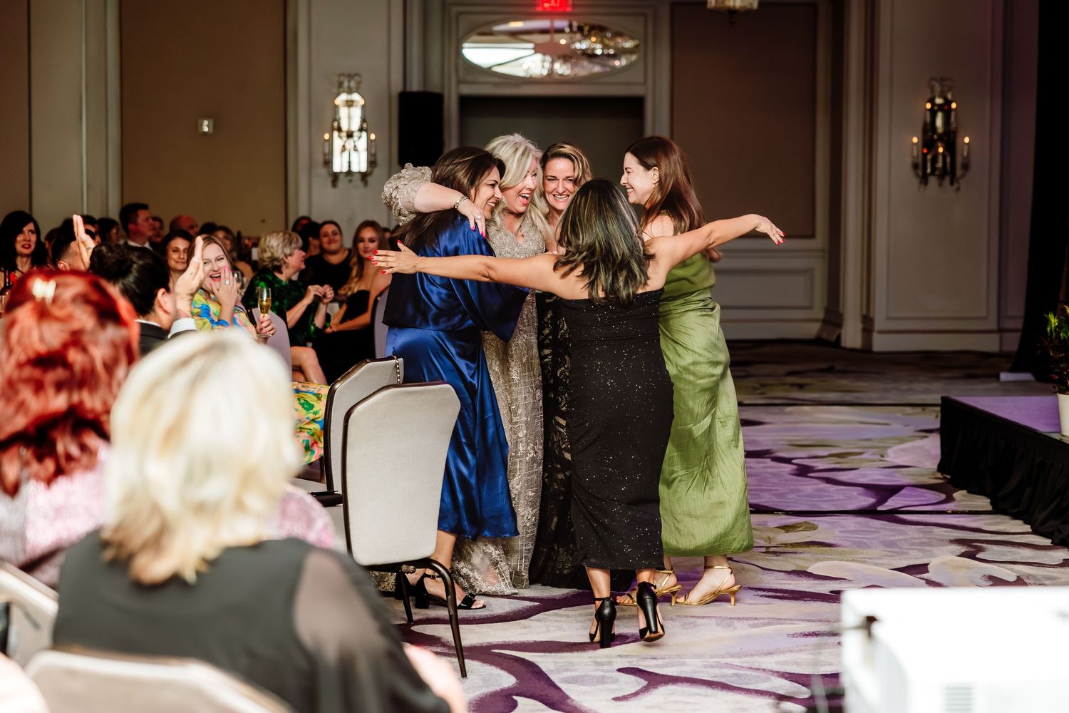 Group of elegantly dressed women embracing on a runway at a formal banquet event.