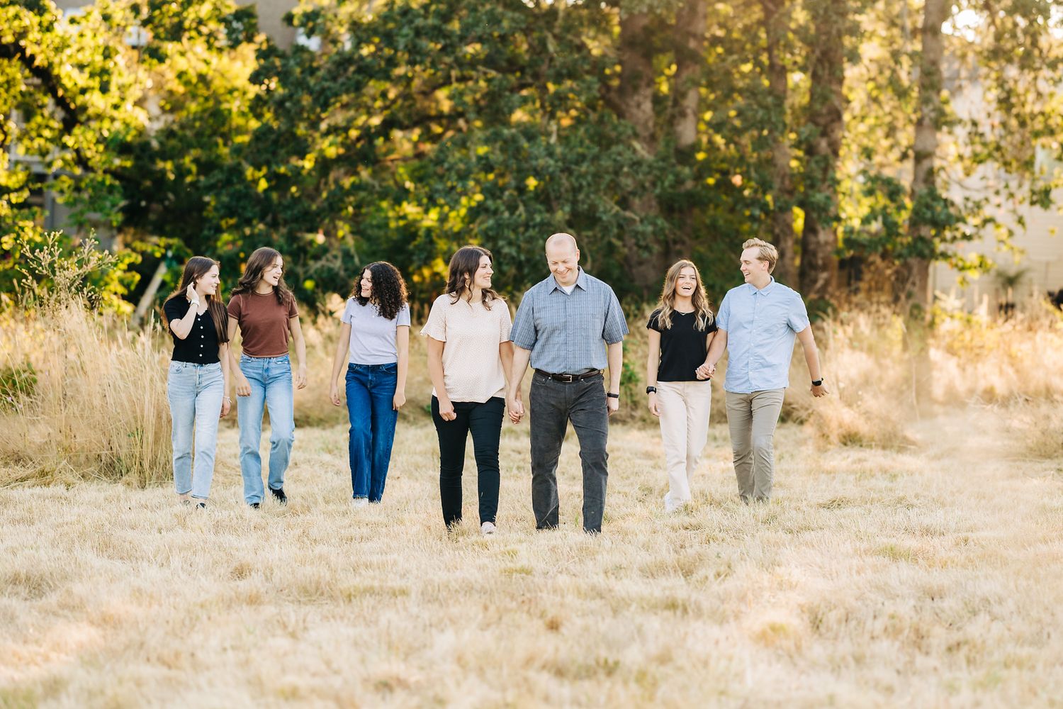 Family of six walking hand-in-hand through golden meadow with lush green trees in warm sunset light.