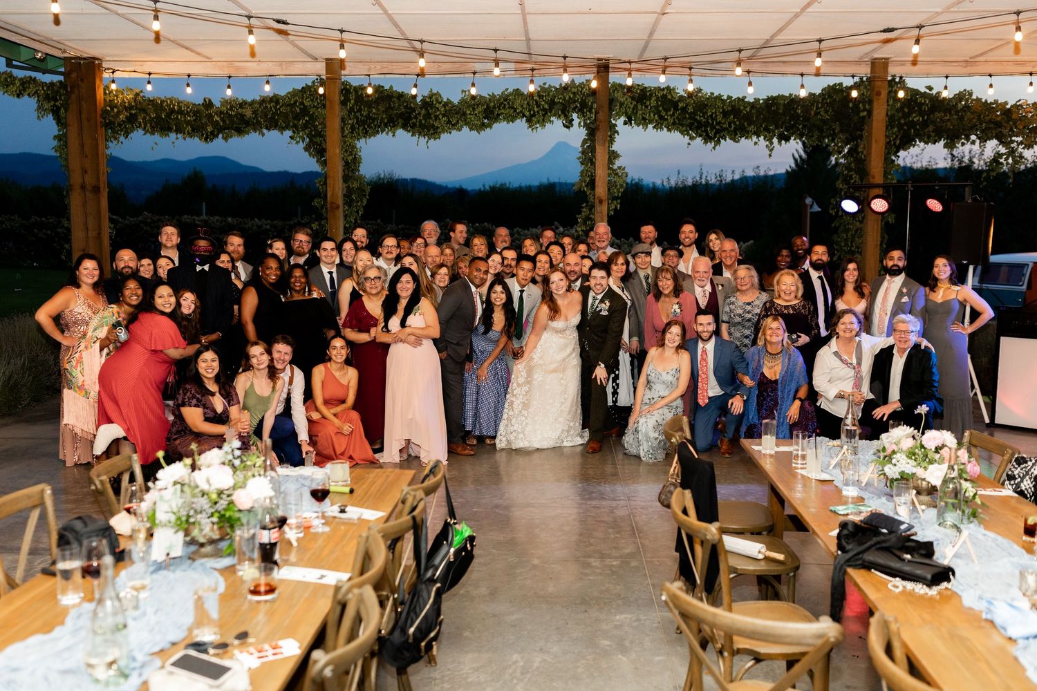 A large wedding party poses together for a group photo under string lights at an outdoor reception venue.
