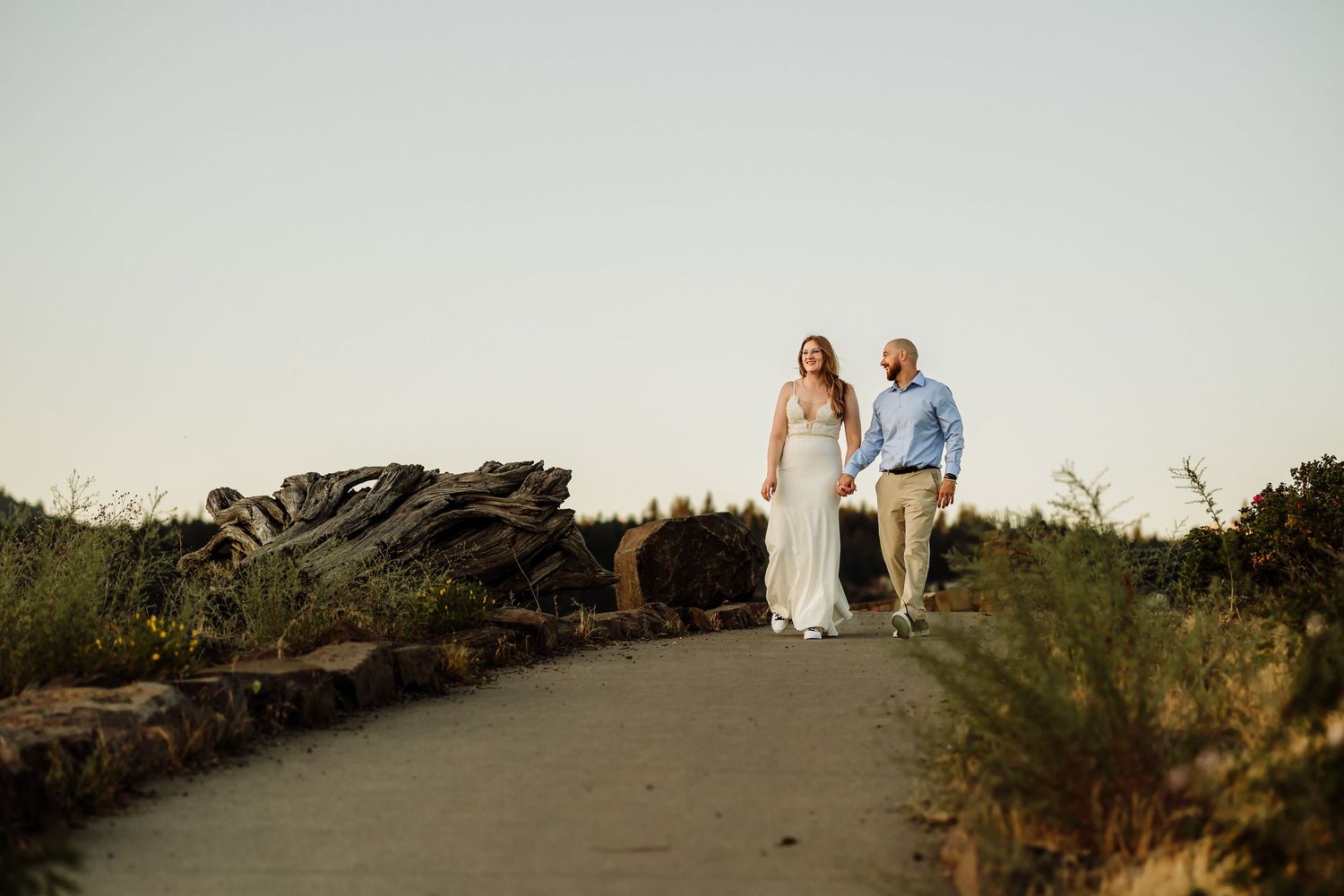 Couple holds hands walking down a sunlit path surrounded by coastal greenery during golden hour engagement session.