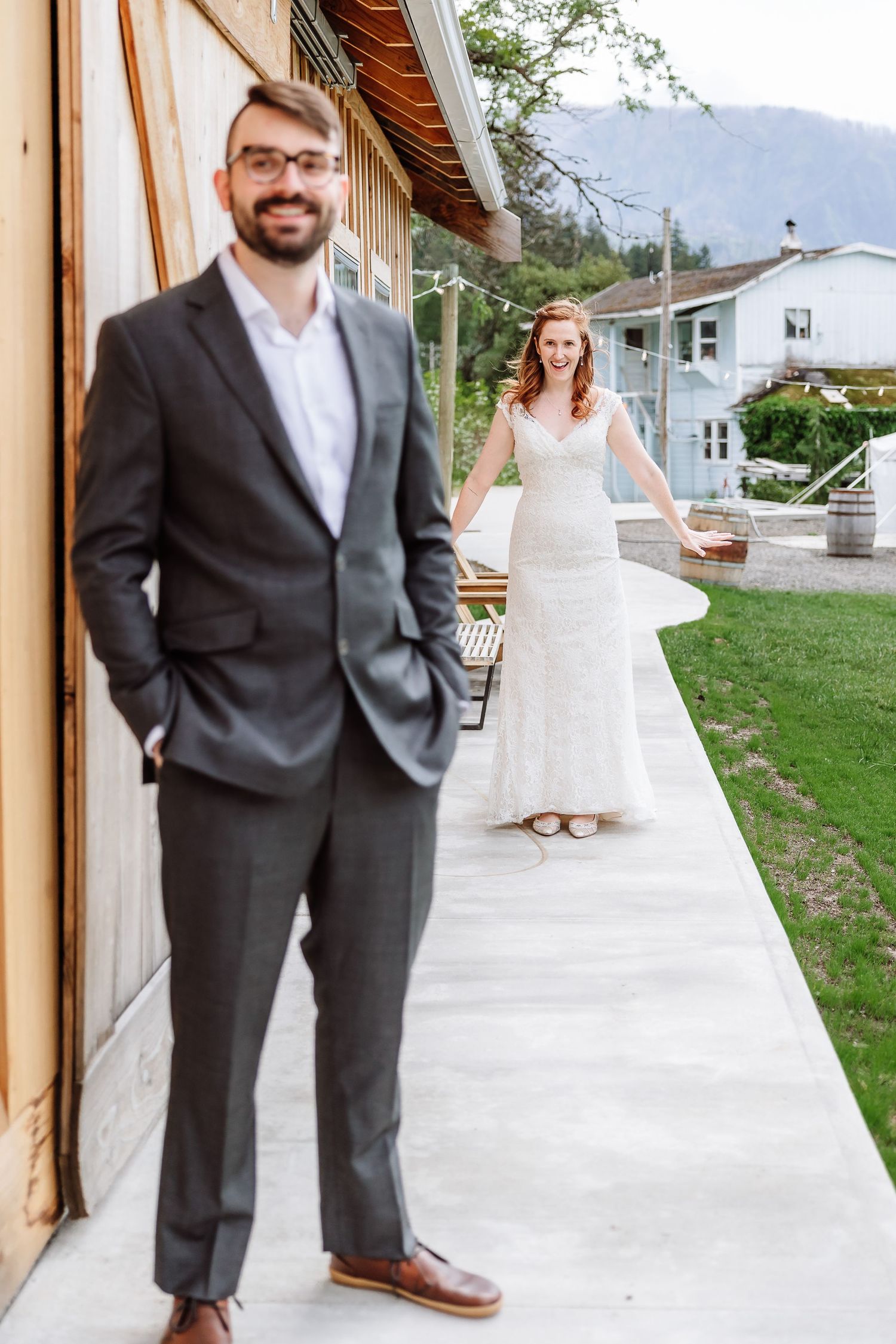 Groom in gray suit stands in foreground as bride in lace gown approaches smiling.