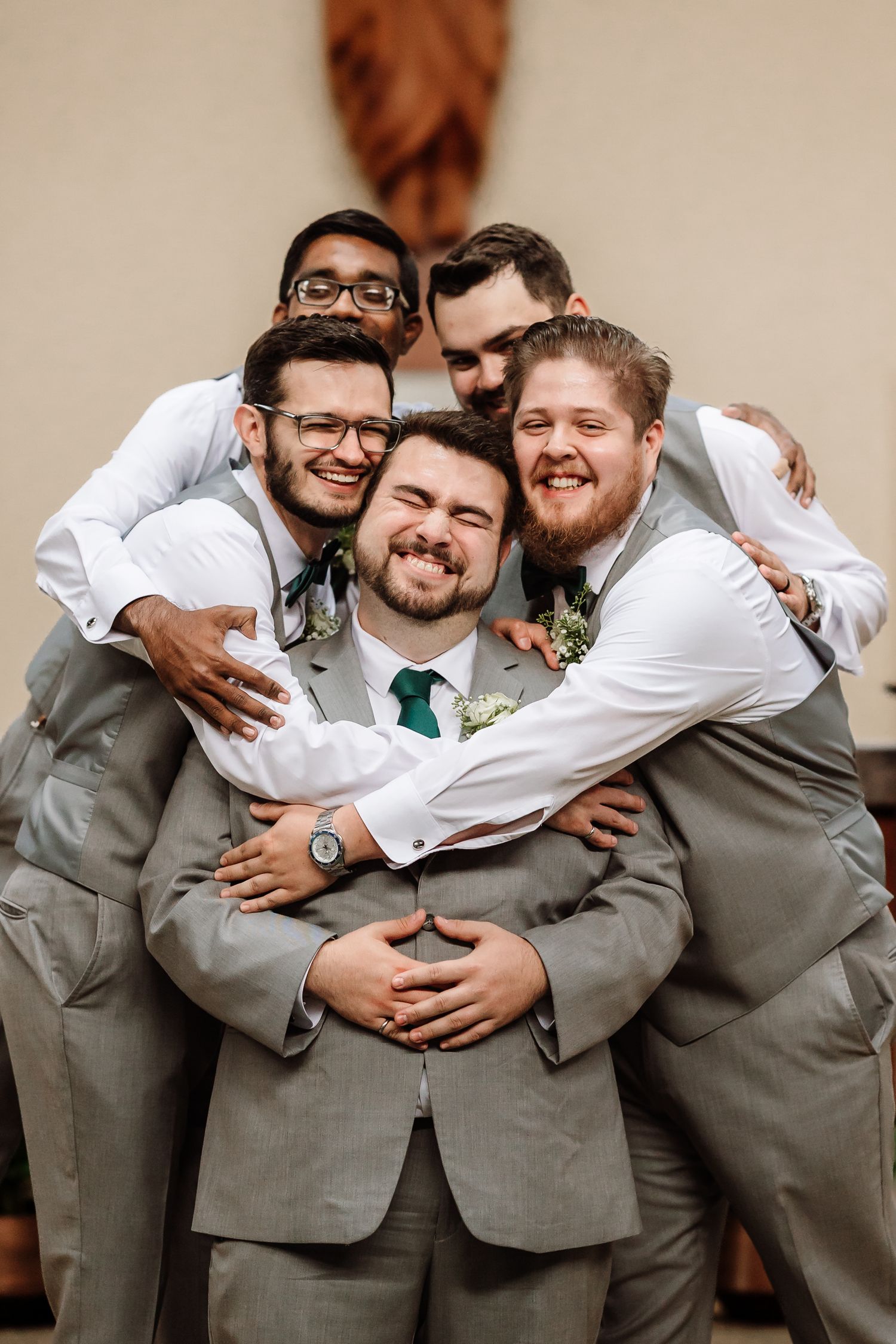 A groom in gray suit is joyfully hugged by four smiling groomsmen in a playful wedding photo.