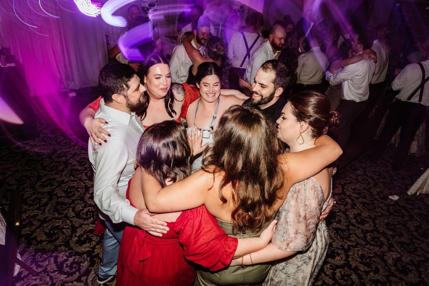 Wedding guests huddle together in a joyful group hug on the dance floor under purple lights.