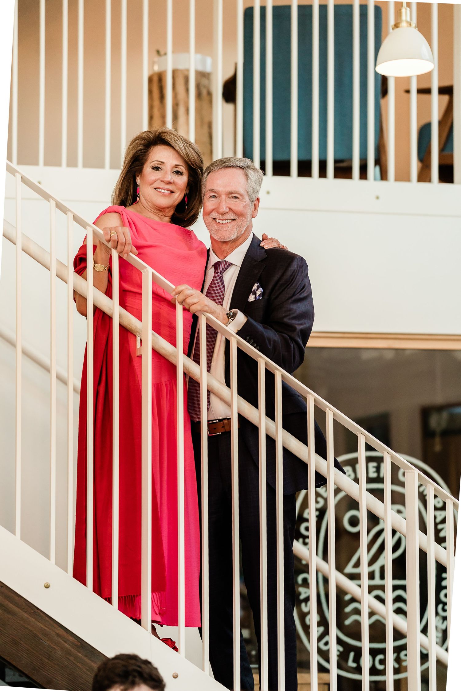 Elegant couple in formal attire smiles together on white staircase at wedding reception venue.