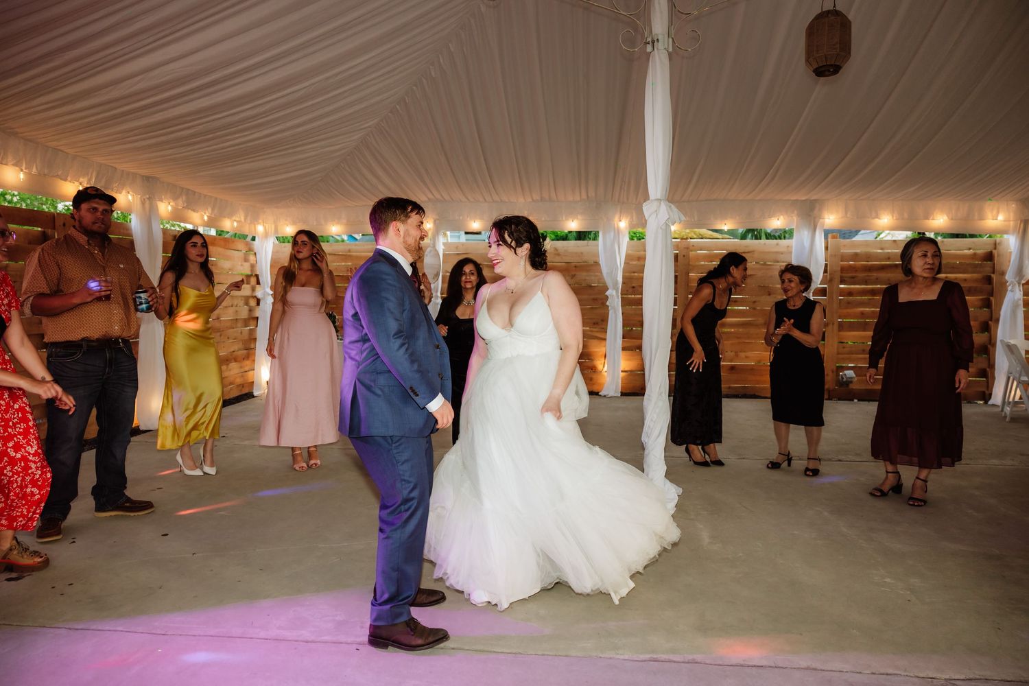 Bride and groom dance together surrounded by guests on a pink-lit dance floor at wedding reception.
