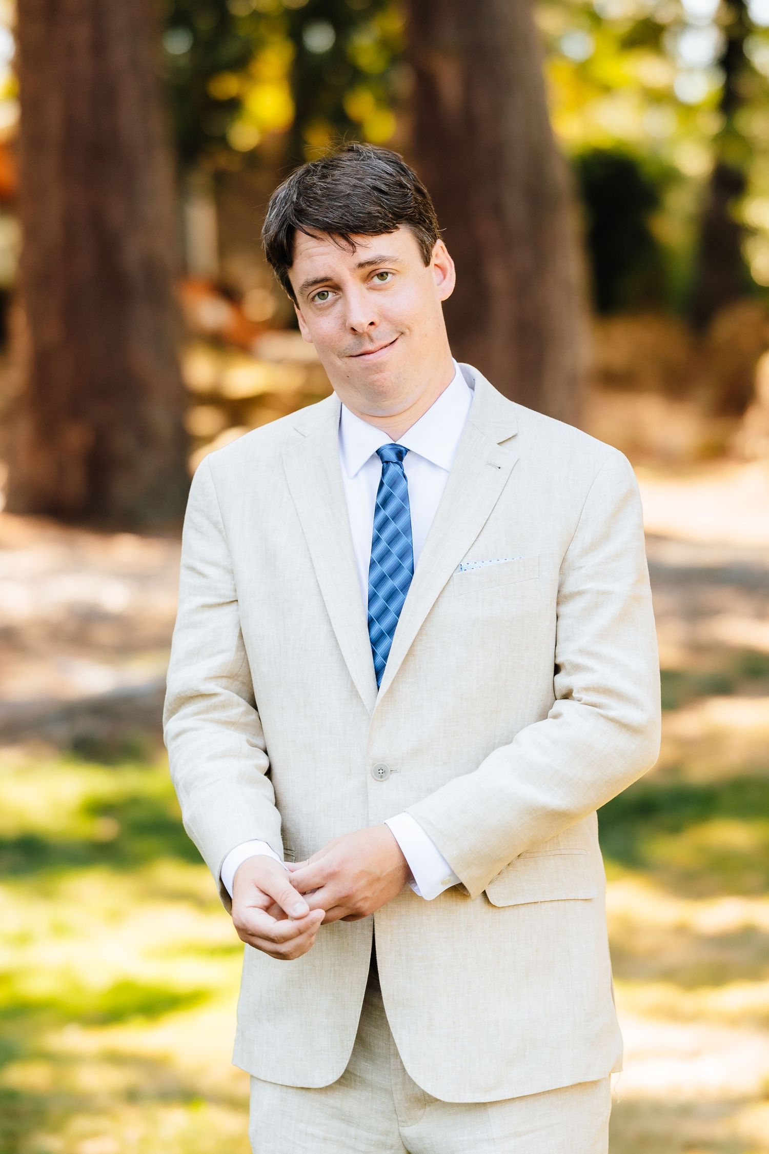 Handsome groom in a beige suit and blue tie smiling confidently outdoors among tall trees on his wedding day.